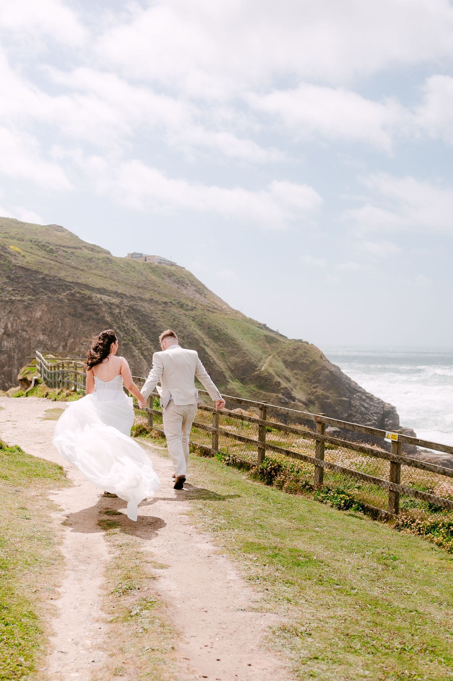 Bride and groom run hand in hand along a scenic coastal cliff path on their wedding day.