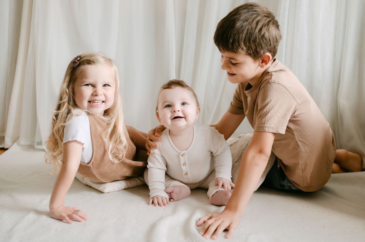 Three young siblings sitting together on a white blanket, two toddlers and a baby smiling in a bright studio setting.
