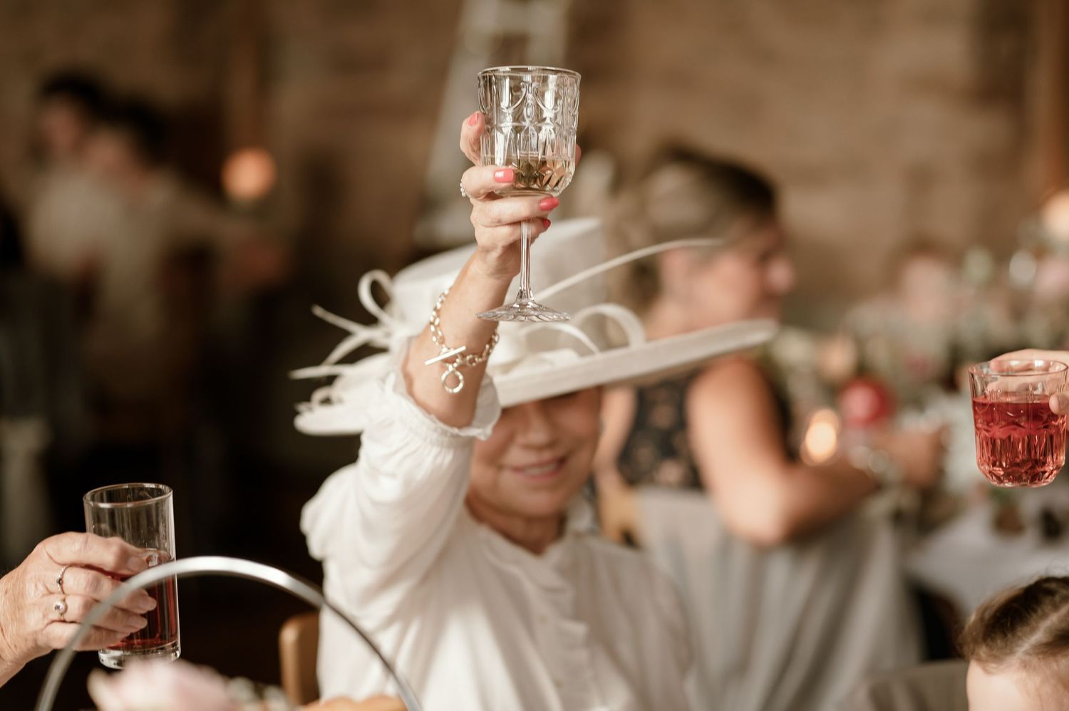 Wedding guest in white hat raises a crystal glass in a toast at elegant indoor wedding reception.