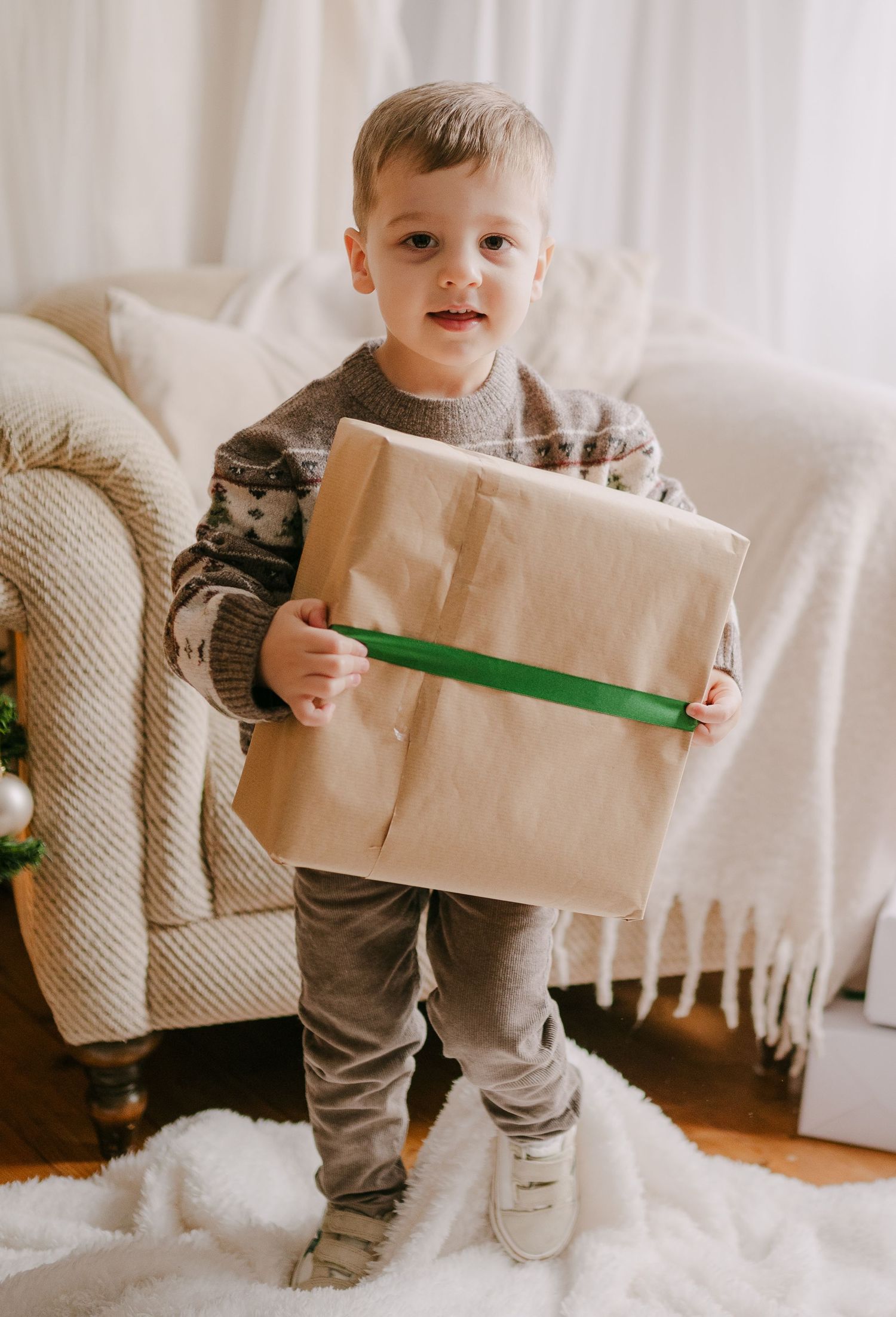 Happy toddler boy in a Christmas sweater holding a large brown kraft paper gift with green ribbon.