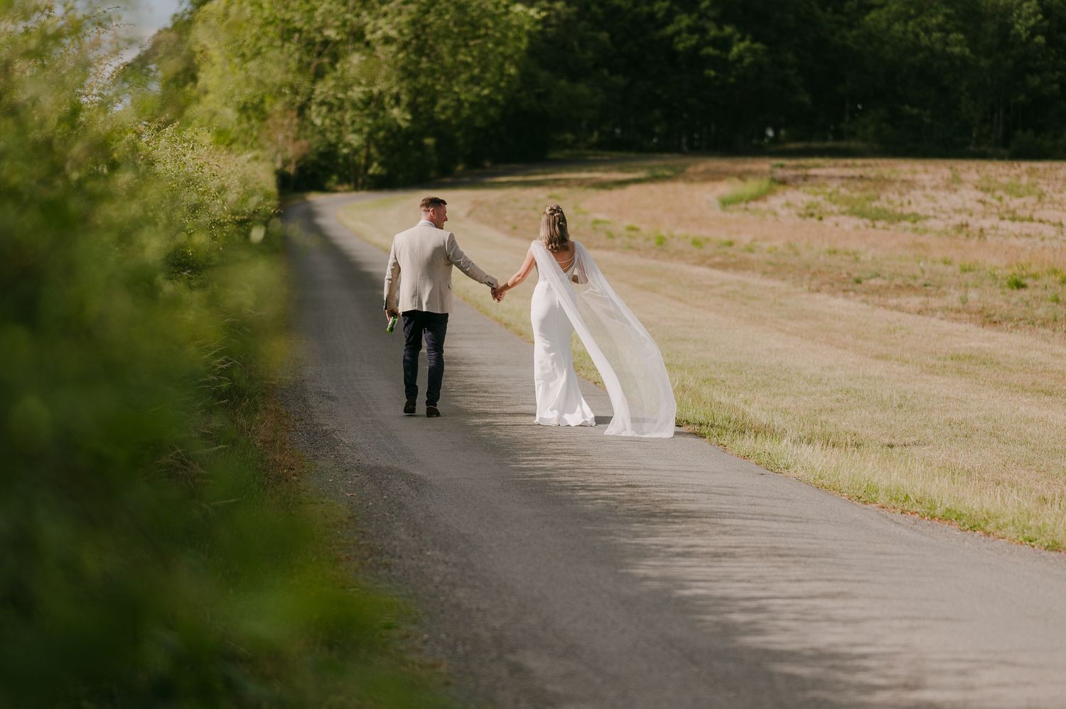 Bride and groom walk hand in hand down a sunlit country road with a flowing white cape veil.