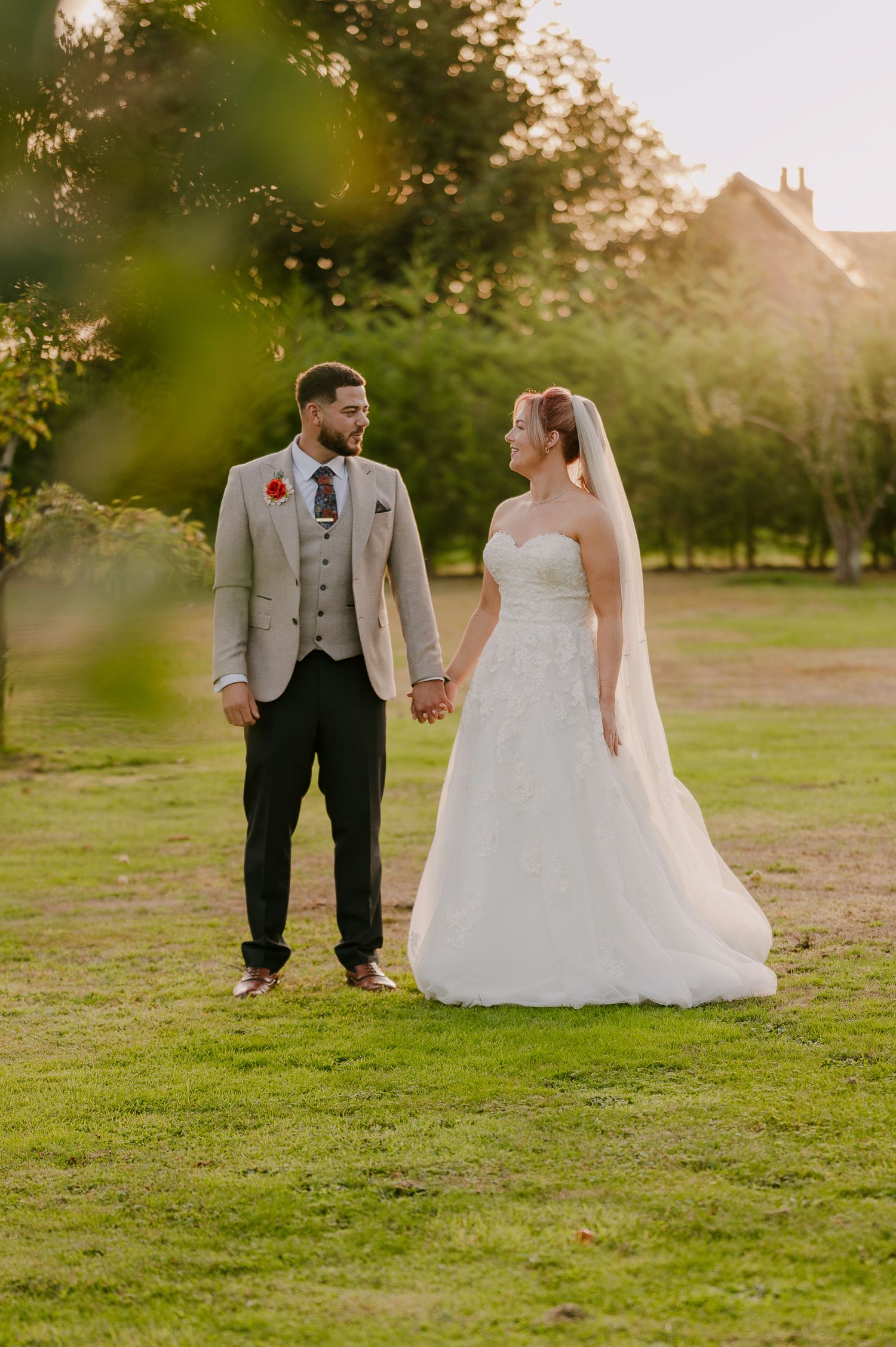 Bride and groom hold hands in sunlit garden, groom in grey suit with red boutonniere, bride in lace gown.