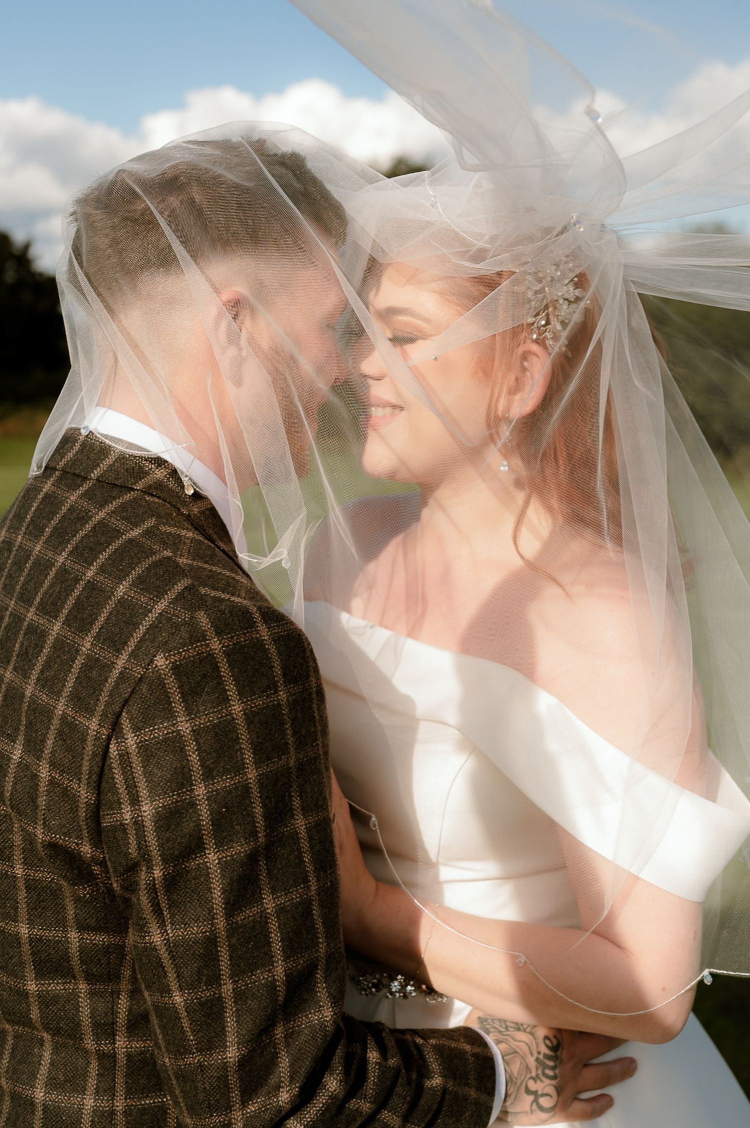 Bride and groom share an intimate moment under a flowing veil on their outdoor wedding day.