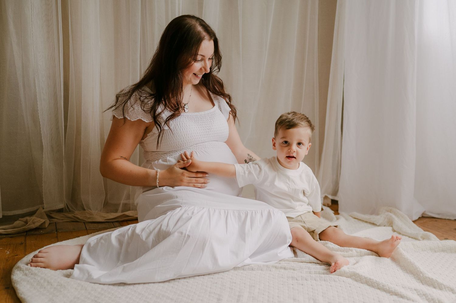 Pregnant mother in white dress sitting with toddler son during a maternity photo session indoors.