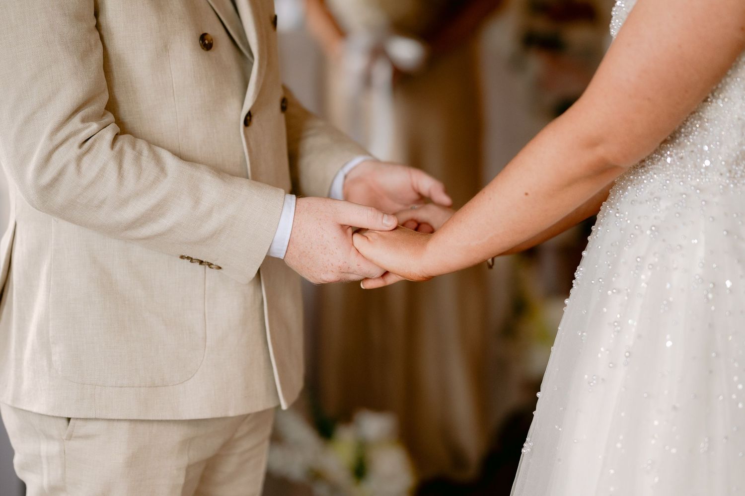 Bride and groom holding hands during wedding ceremony, groom wearing a beige suit and bride in a beaded gown.