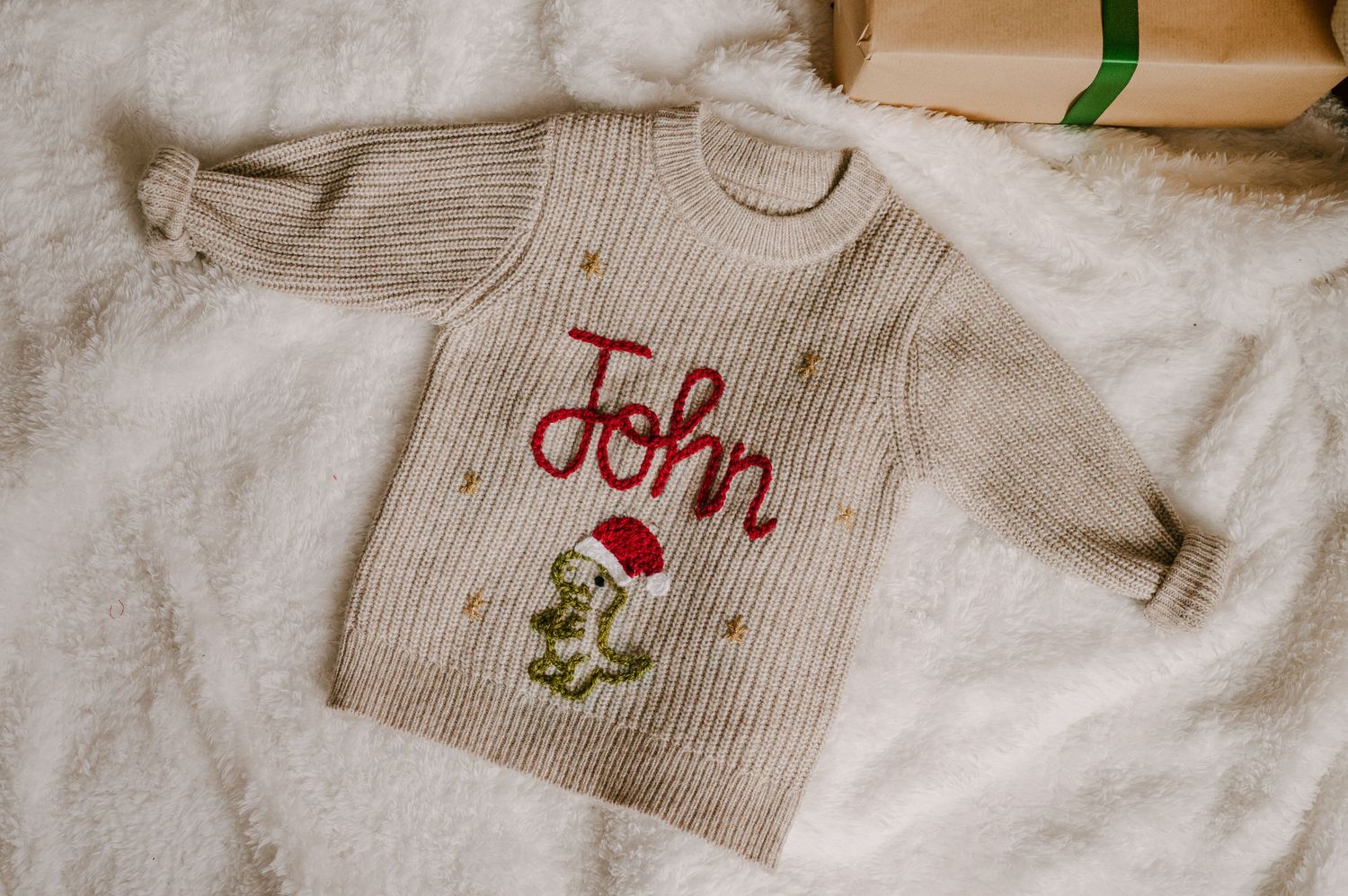 Personalized beige knit Christmas sweater with John embroidered in red and a festive dinosaur wearing a Santa hat.