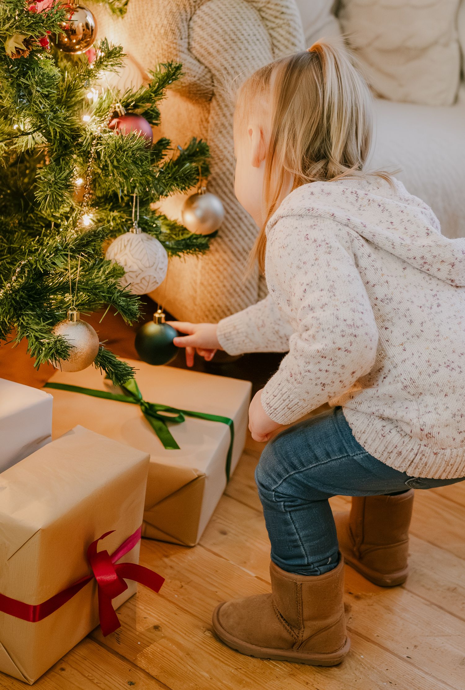 Little girl in cream sweater and boots crouching to touch ornaments under a decorated Christmas tree.
