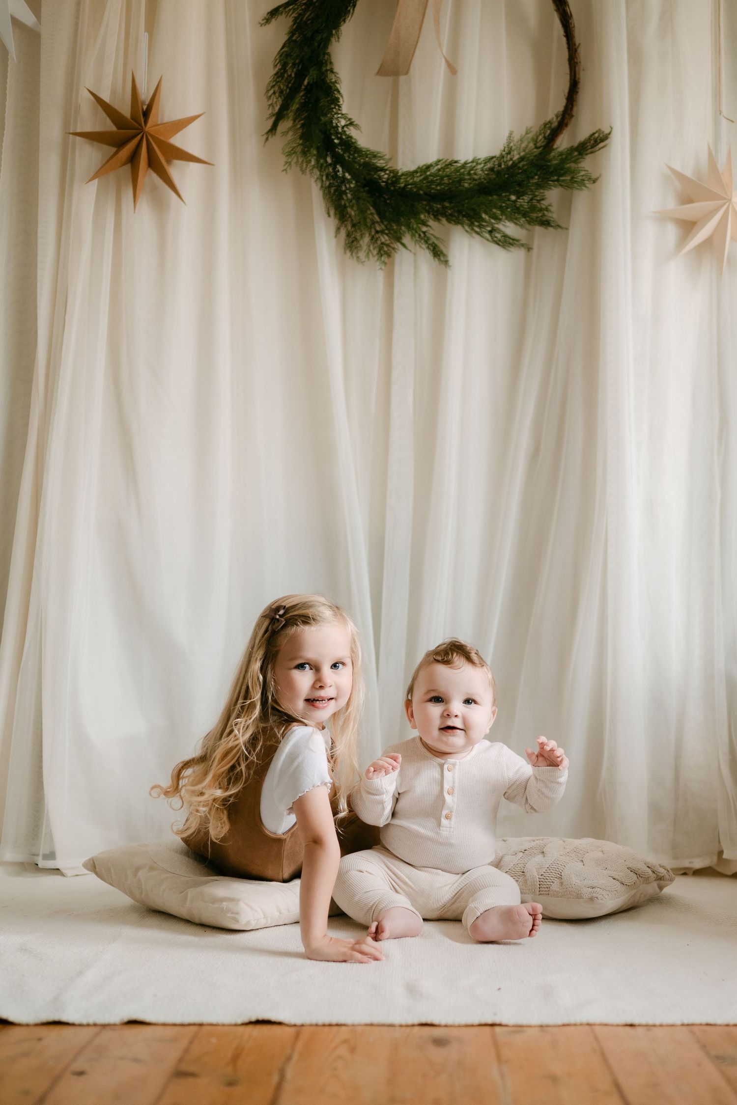 Two young children sit on pillows before a festive backdrop with a wreath and paper stars.