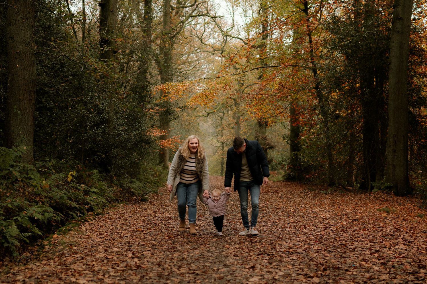 Family of three walking hand-in-hand through an autumn forest path covered in fallen leaves.