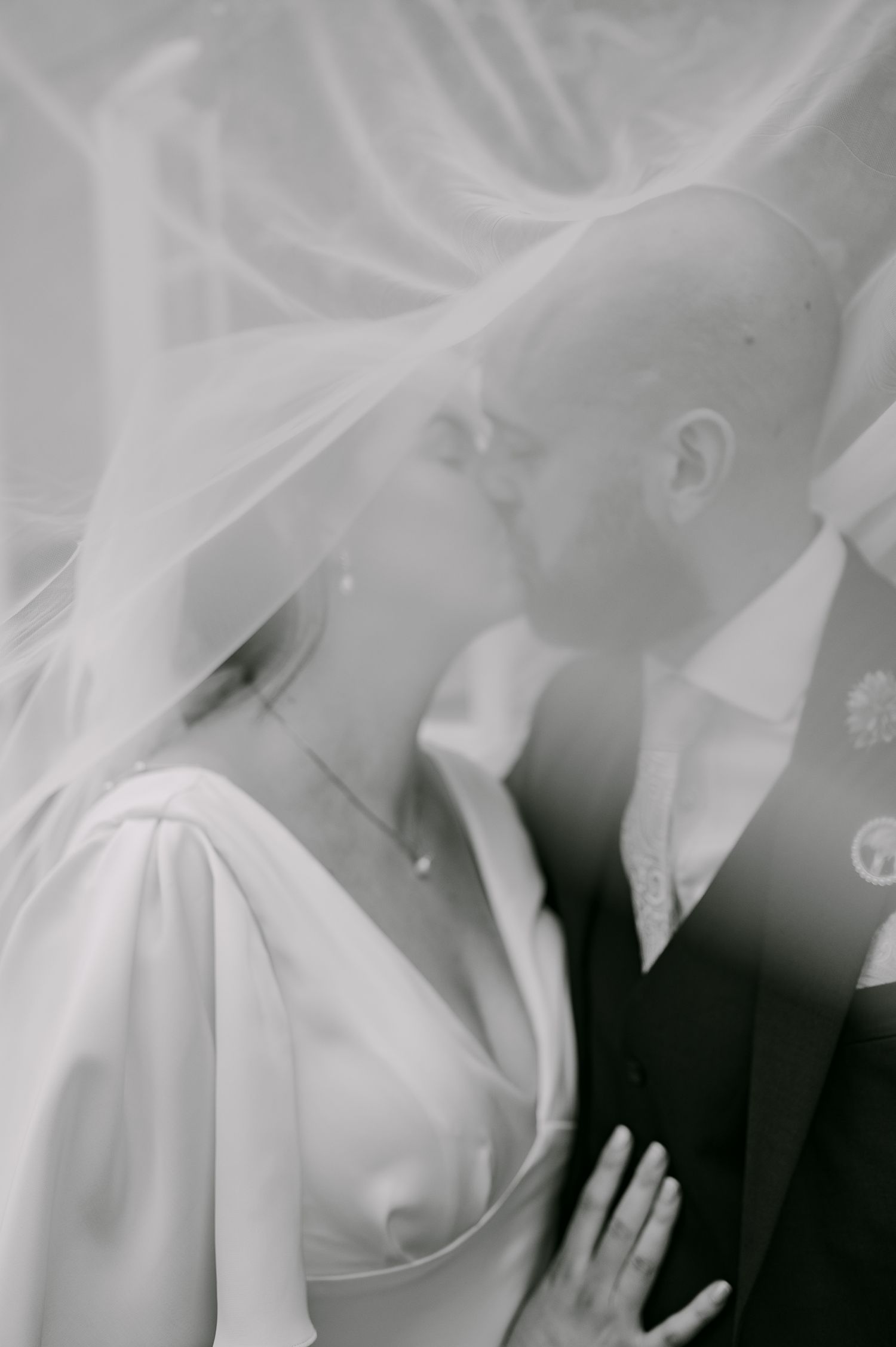 Black and white romantic photo of bride and groom kissing under a flowing wedding veil.
