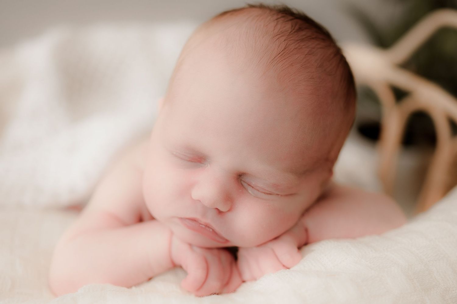 Sleeping newborn baby resting chin on hands in a soft white fluffy basket with warm lighting.