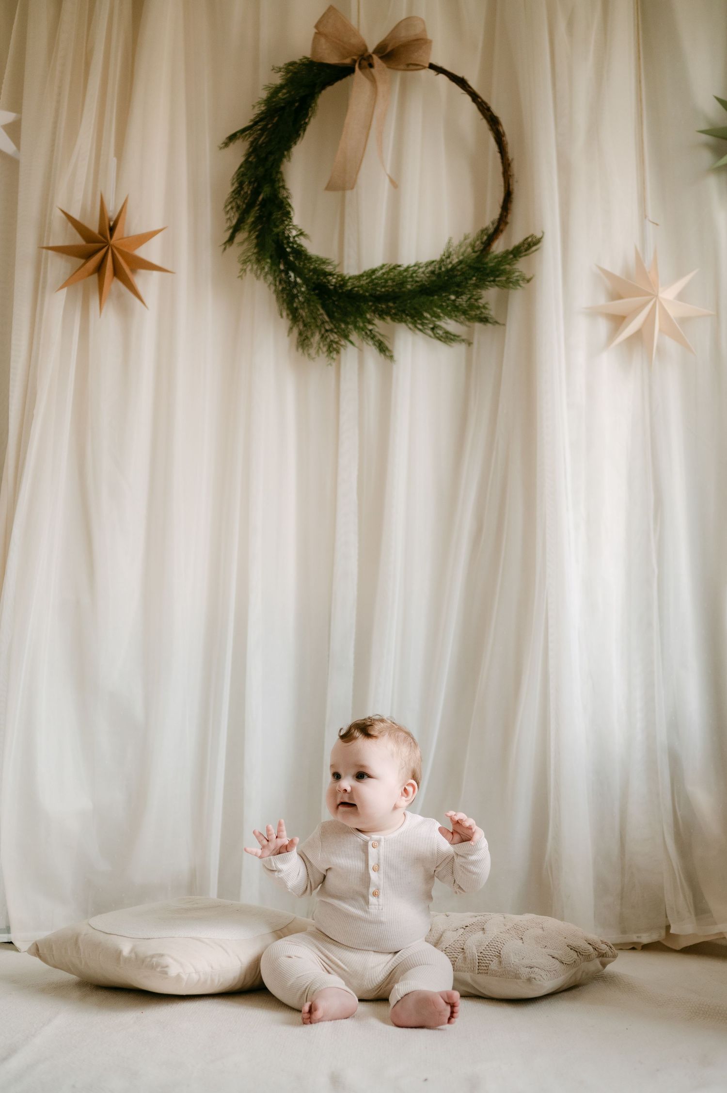 A baby in white outfit sits on pillows before a festive backdrop with a pine wreath, bow, and star decorations.