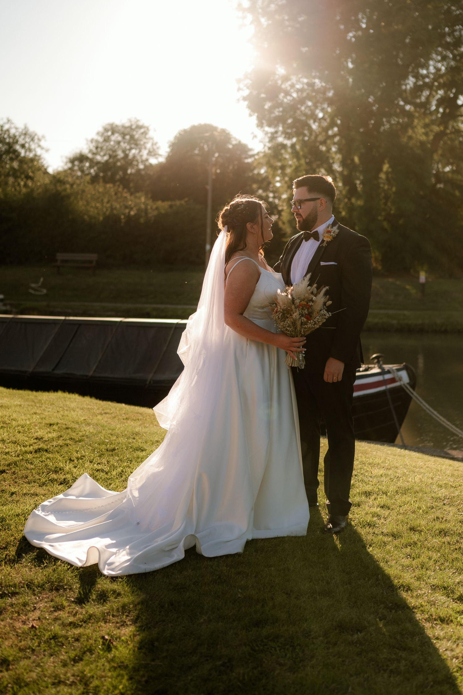 Bride and groom share romantic golden hour moment by riverside, bride in flowing white gown with veil.