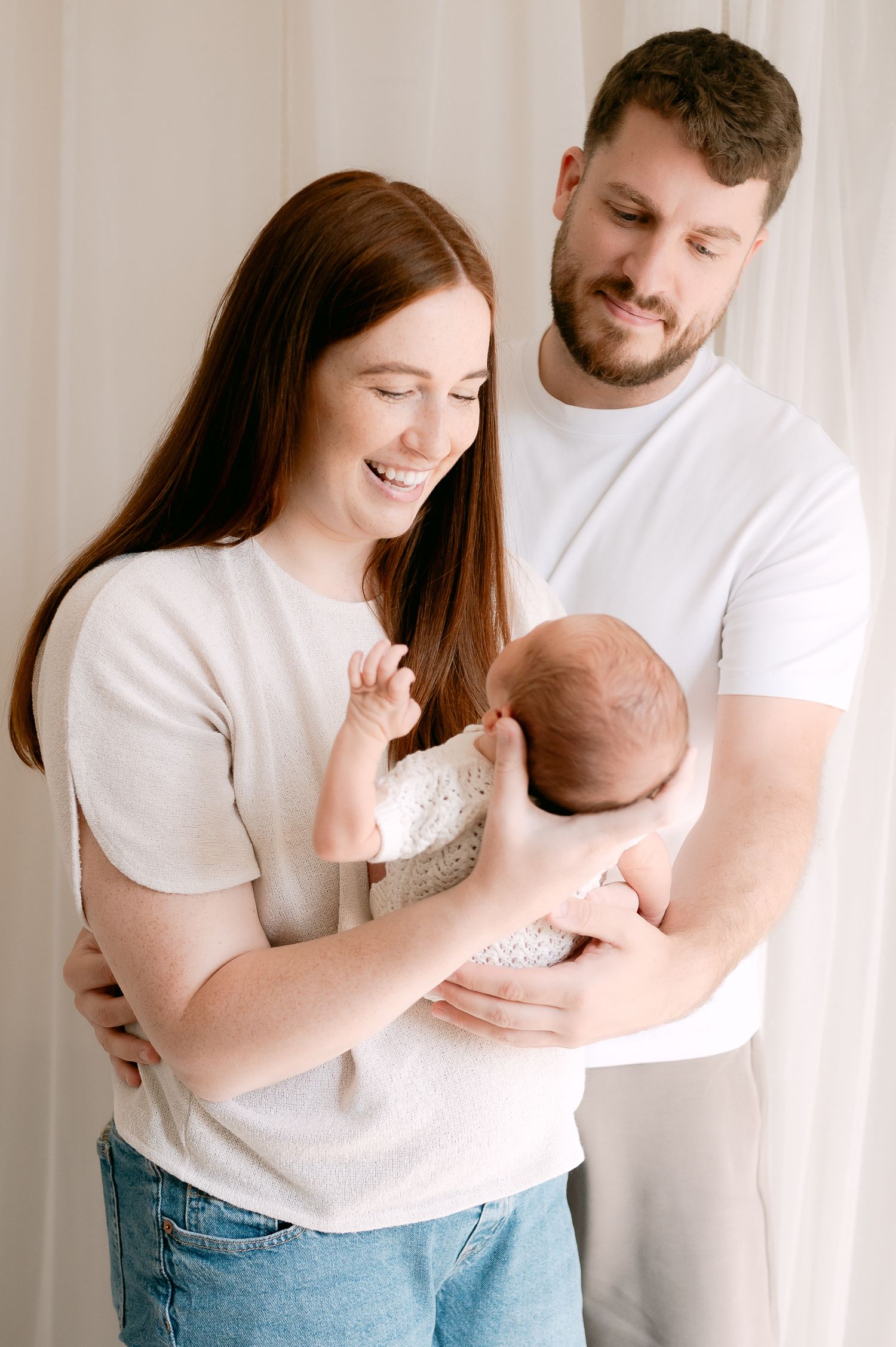 Happy parents smiling and holding their newborn baby together in a bright white room.