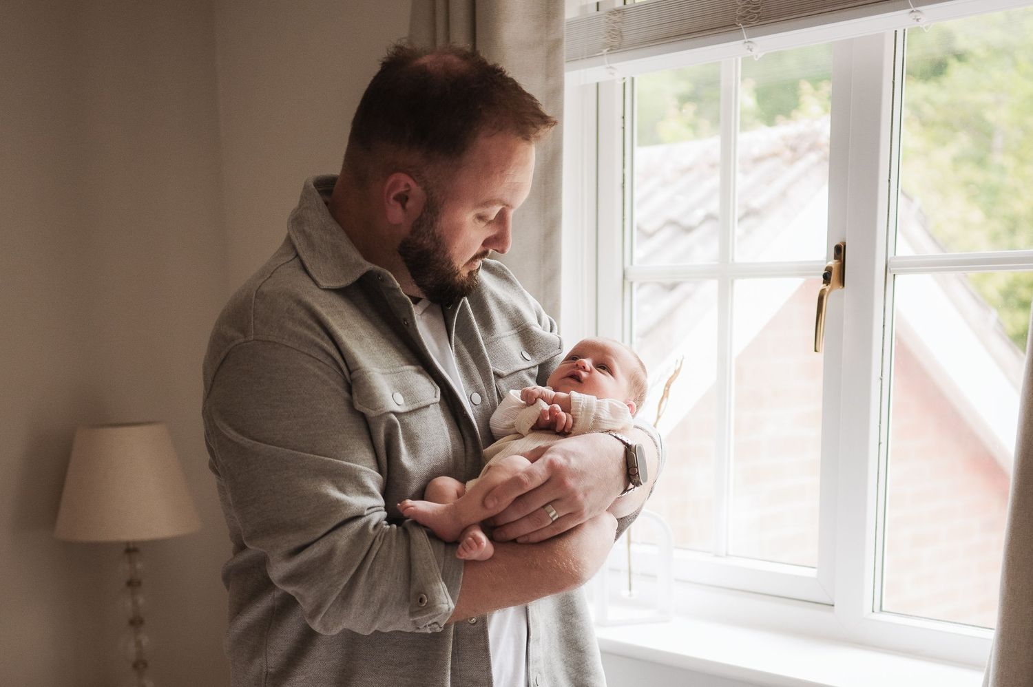 Father standing by window gazing lovingly at newborn baby in arms.