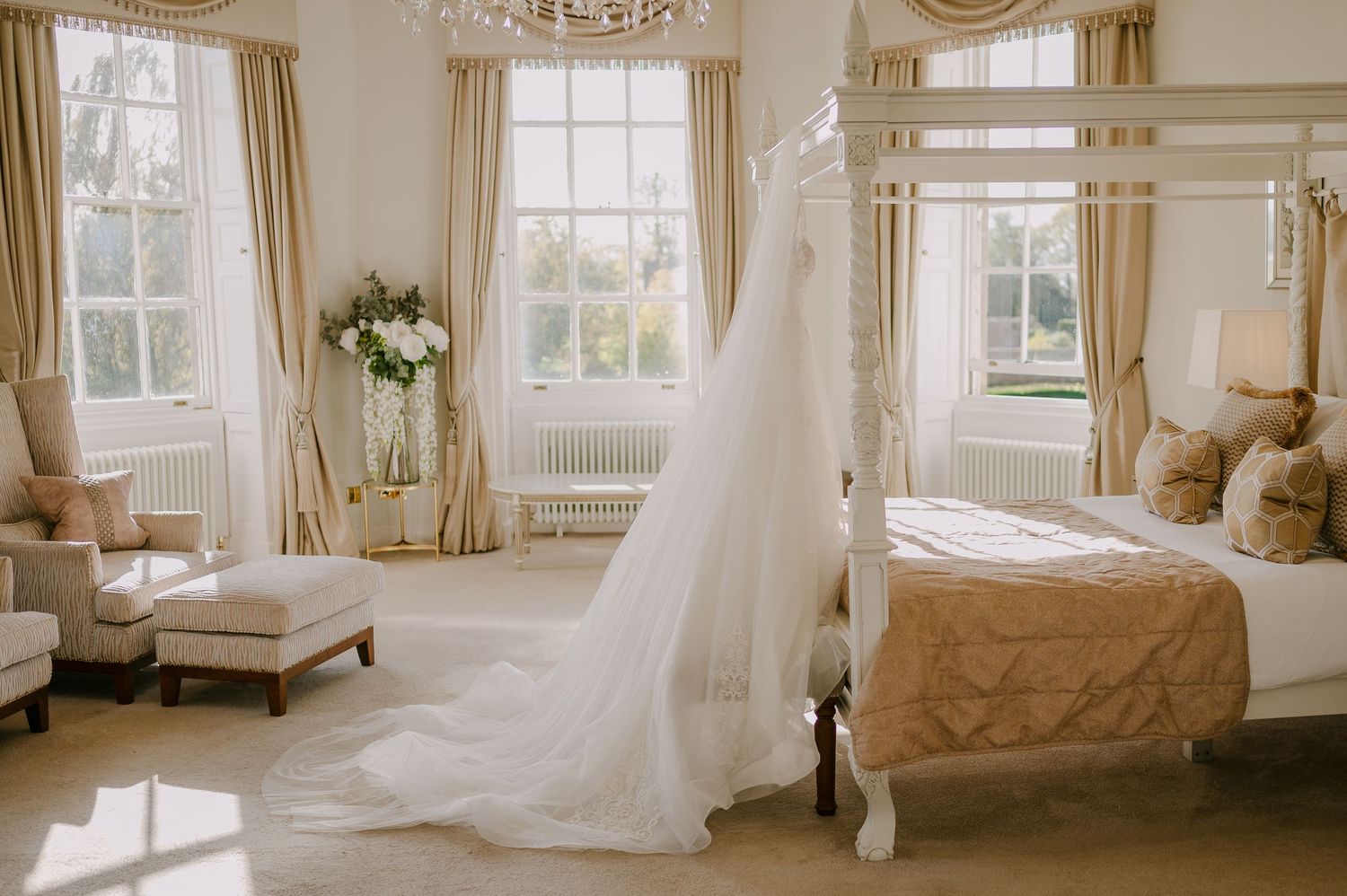 Elegant wedding dress with long veil draped over four-poster bed in sunlit luxury bridal suite.