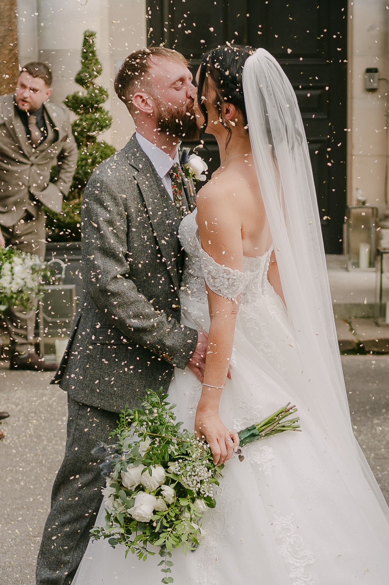 Newlyweds sharing a kiss in a shower of gold confetti outside church, bride holding white rose bouquet.