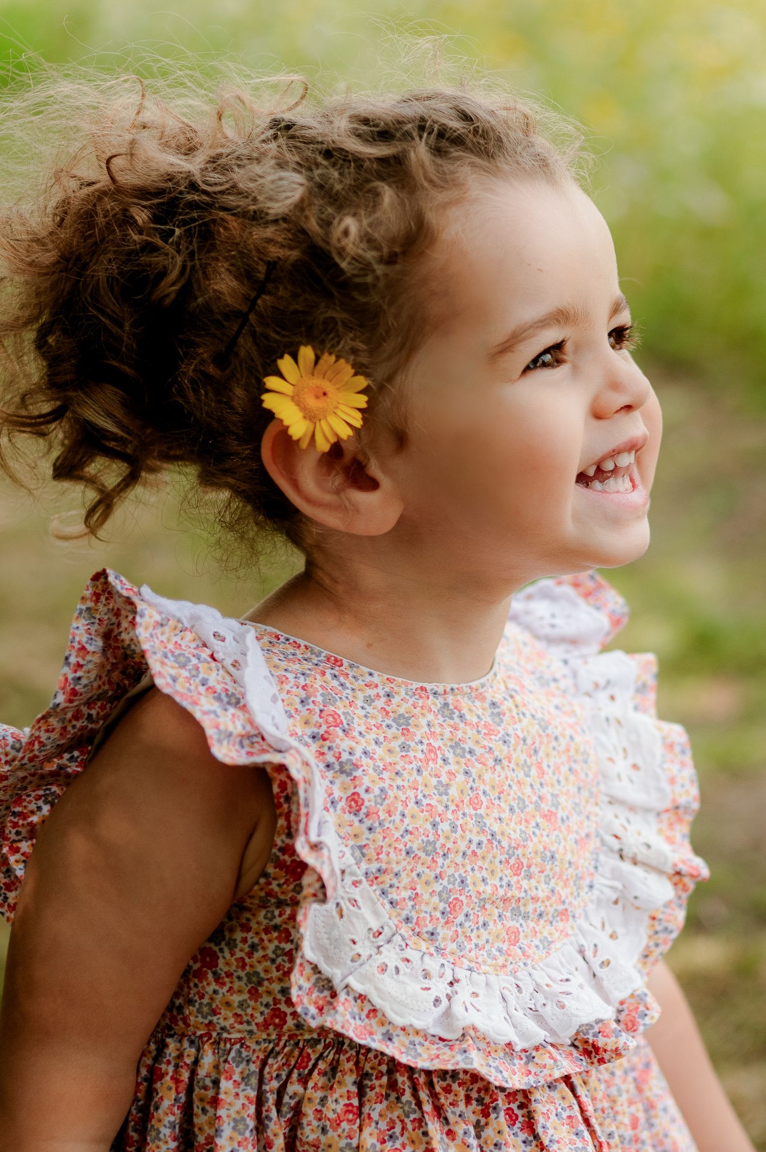 Happy toddler girl with curly hair and yellow flower accessory wearing a floral ruffle dress.