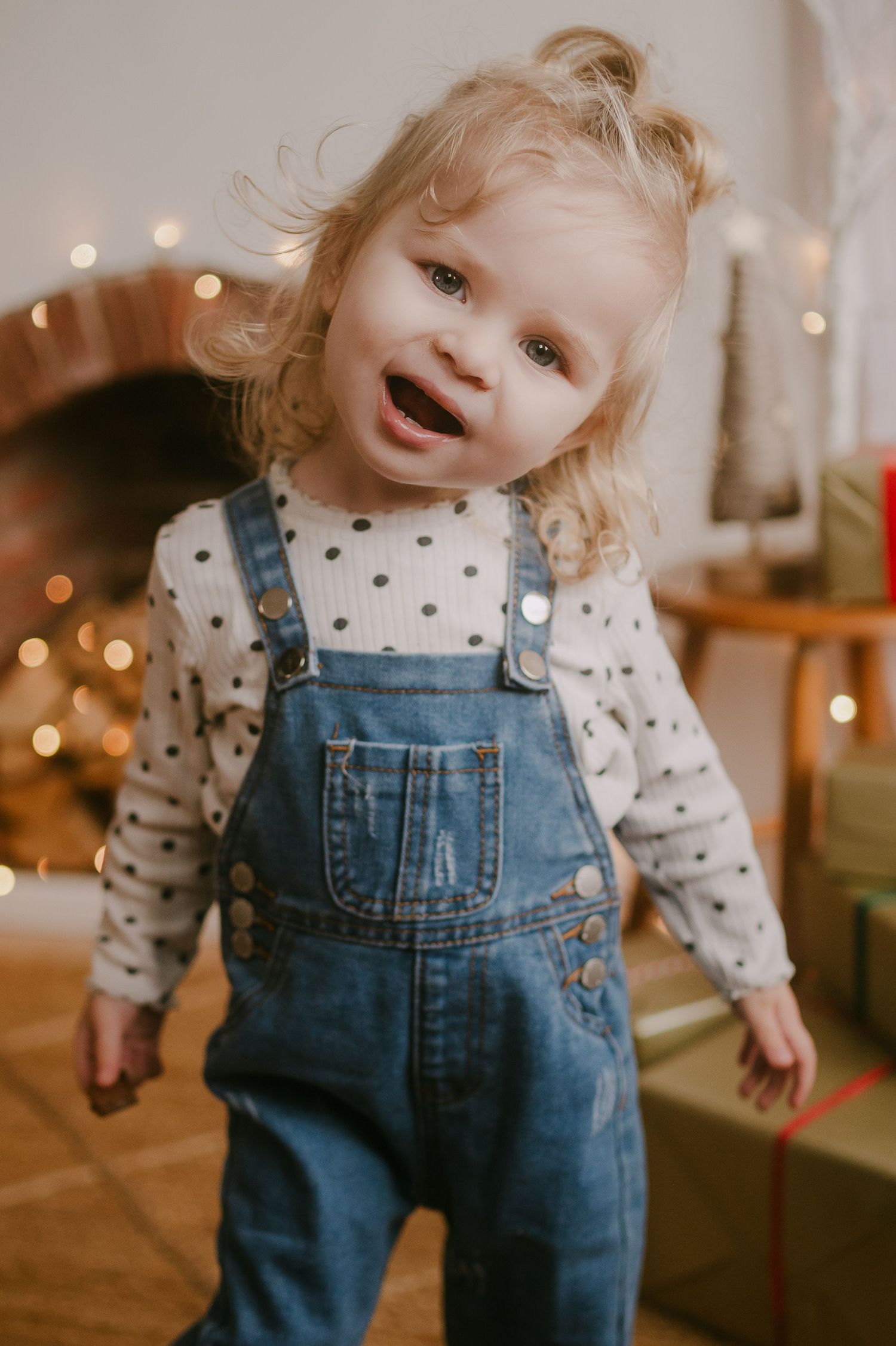 Smiling toddler girl in denim overalls and polka dot shirt near Christmas lights.