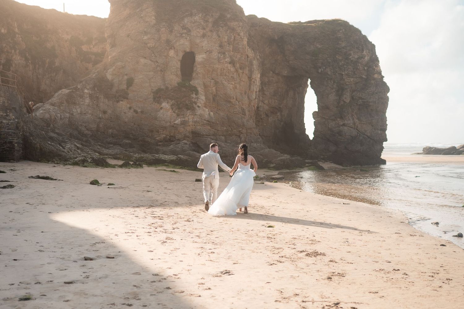 Bride and groom walking hand in hand on a sandy beach with dramatic rock arch formation in golden sunlight.