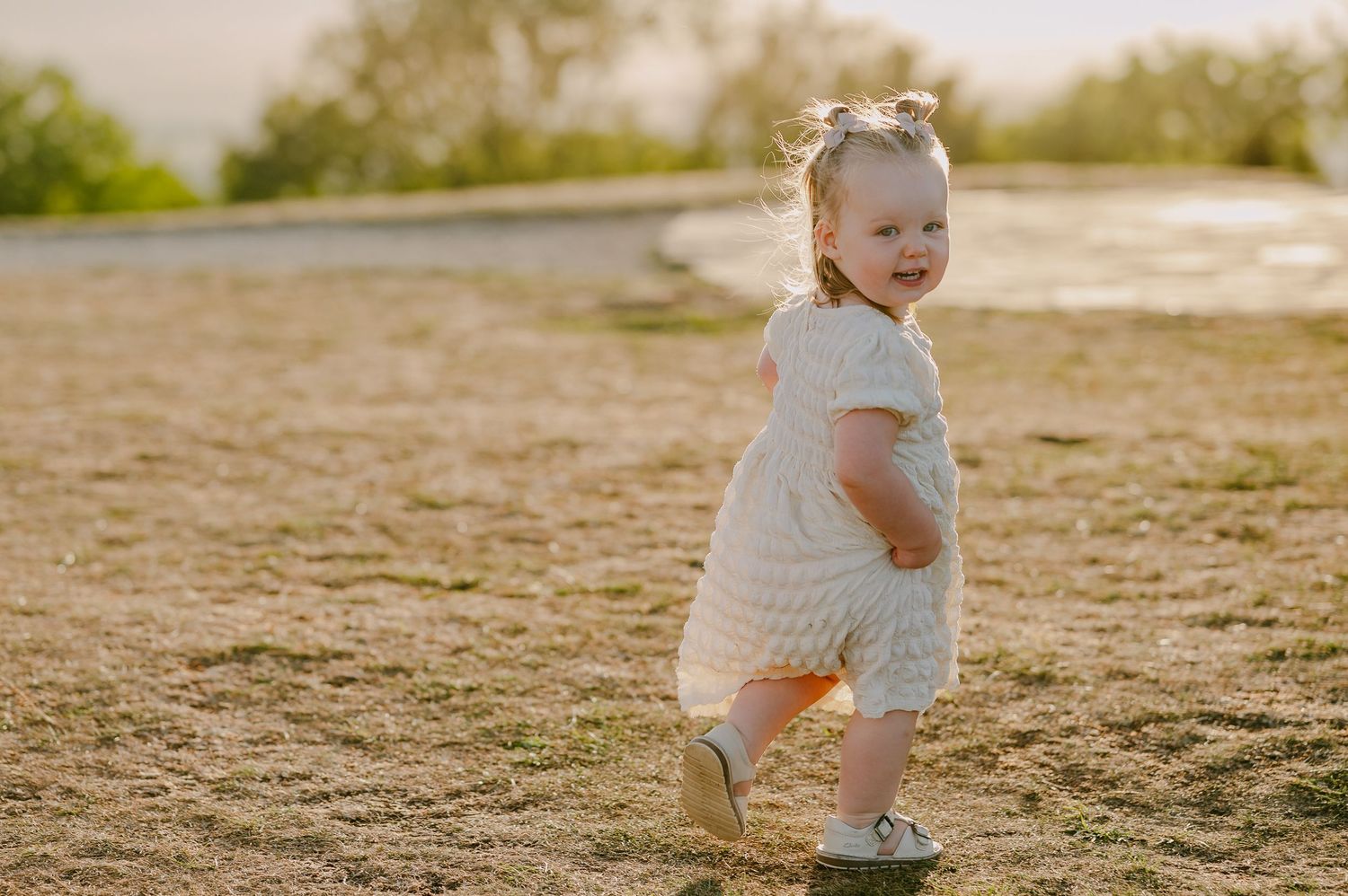 Toddler girl in white dress walking outdoors in golden sunset light.