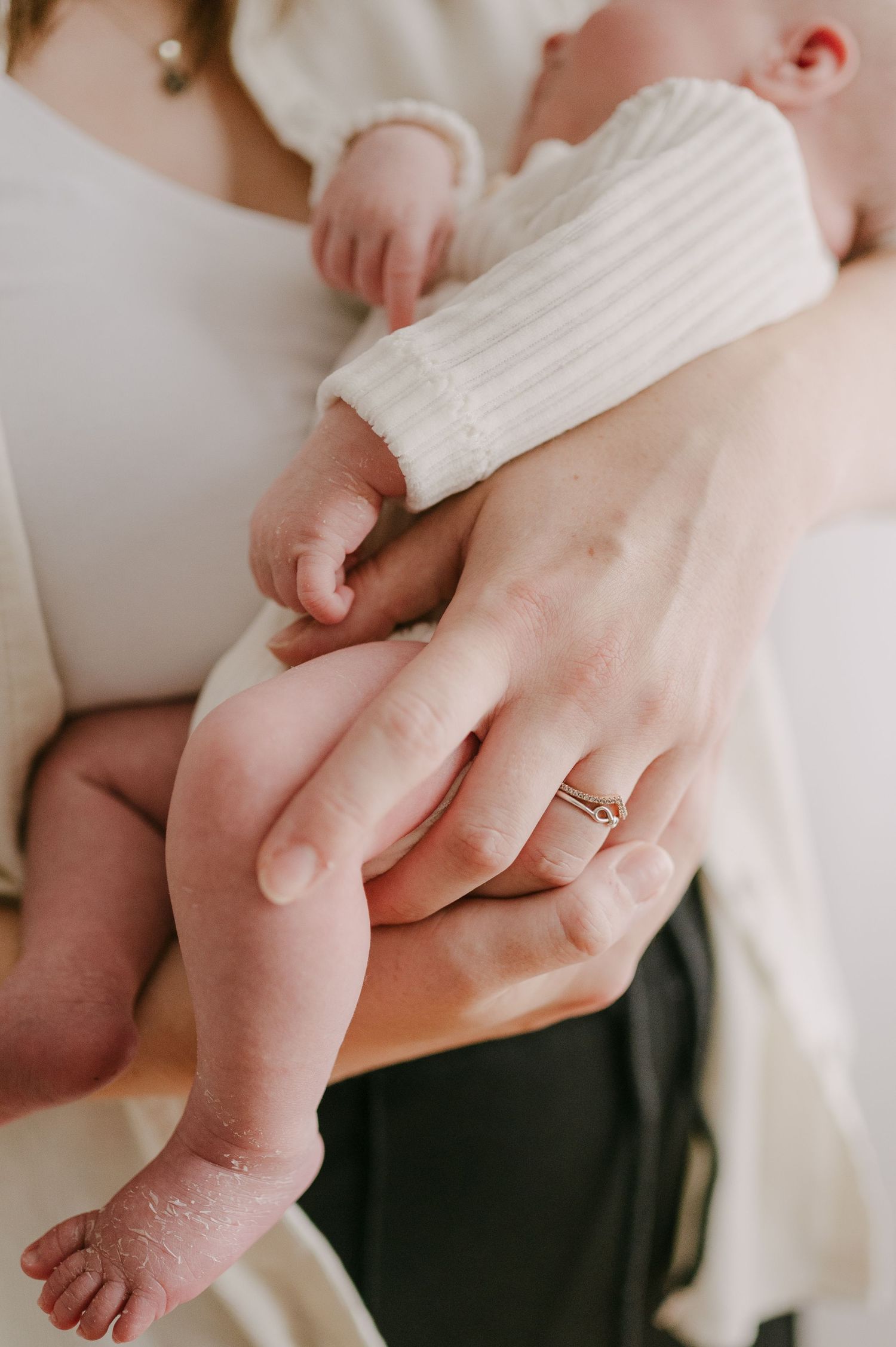 Close-up of mother's hands cradling newborn baby's legs and feet with a delicate ring.