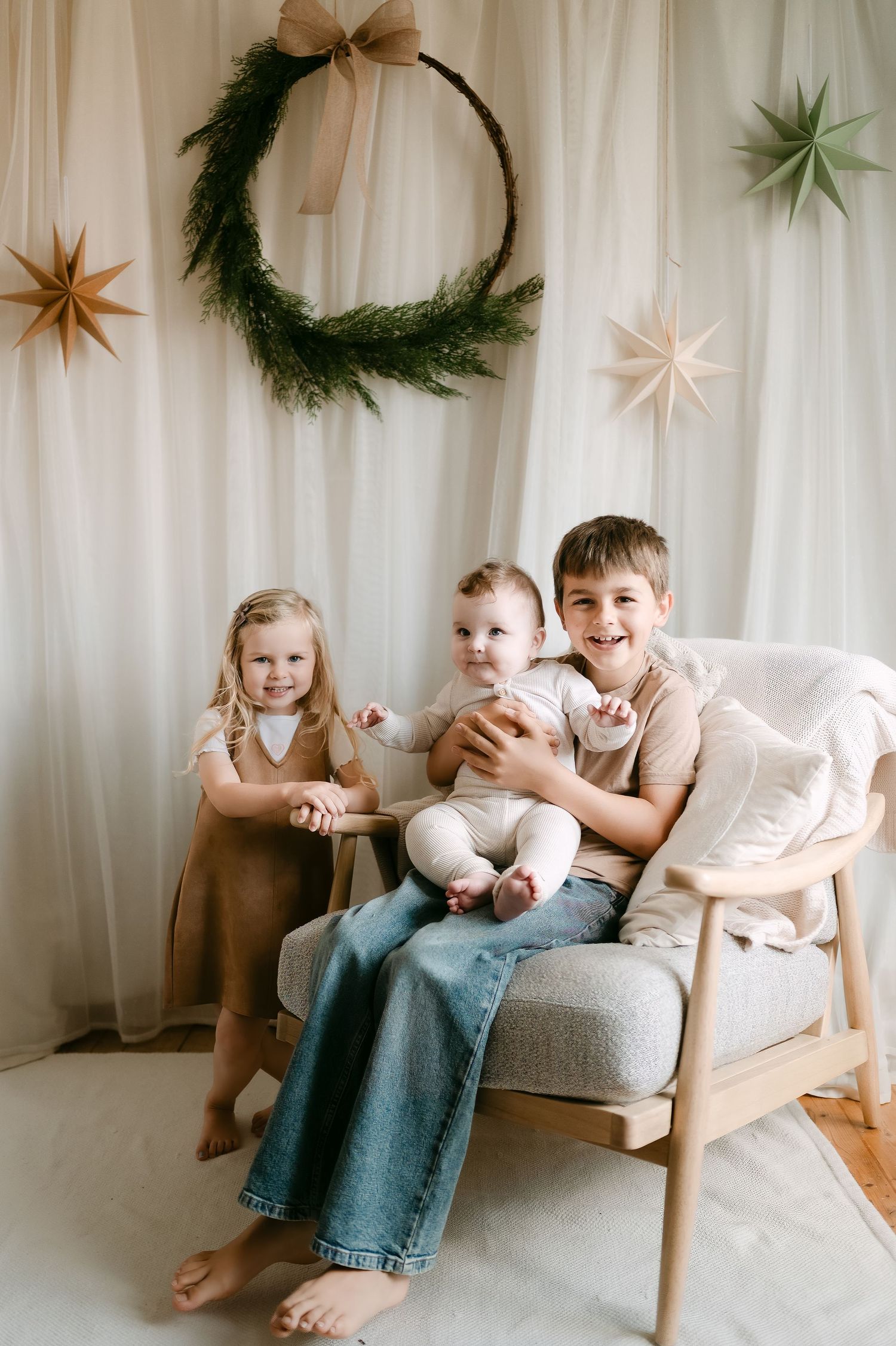 Three children pose for a festive holiday photo with a wreath and star decorations in a bright, neutral-toned studio setting.