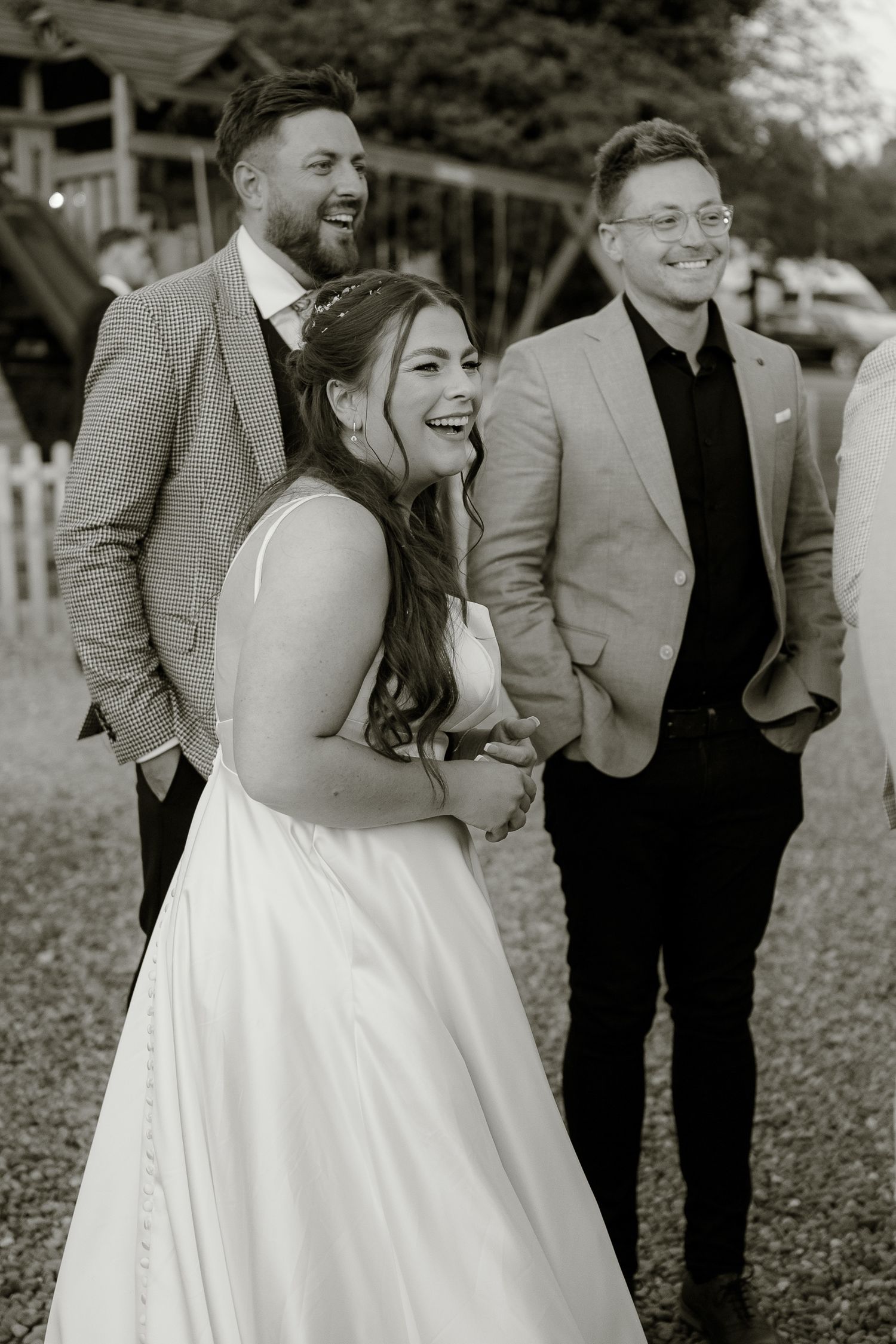 Black and white photo of a laughing bride in white gown with two smiling men in suits at outdoor wedding.