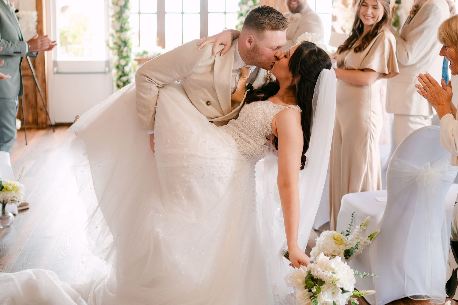 Groom dips bride for romantic kiss during wedding ceremony, bride holding white floral bouquet.
