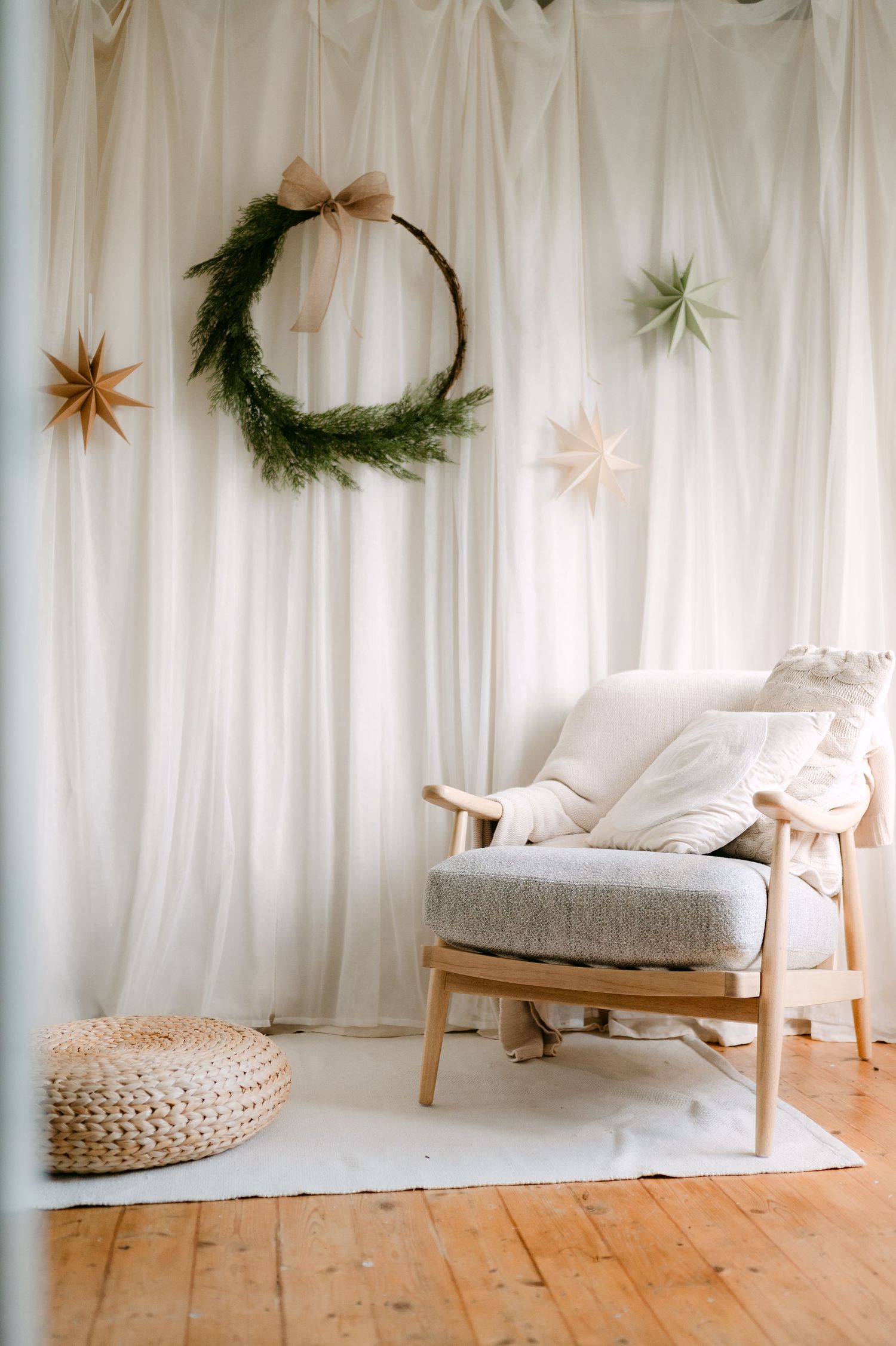 Scandinavian Christmas living room with wooden armchair, evergreen wreath, paper stars, and white draped curtains.
