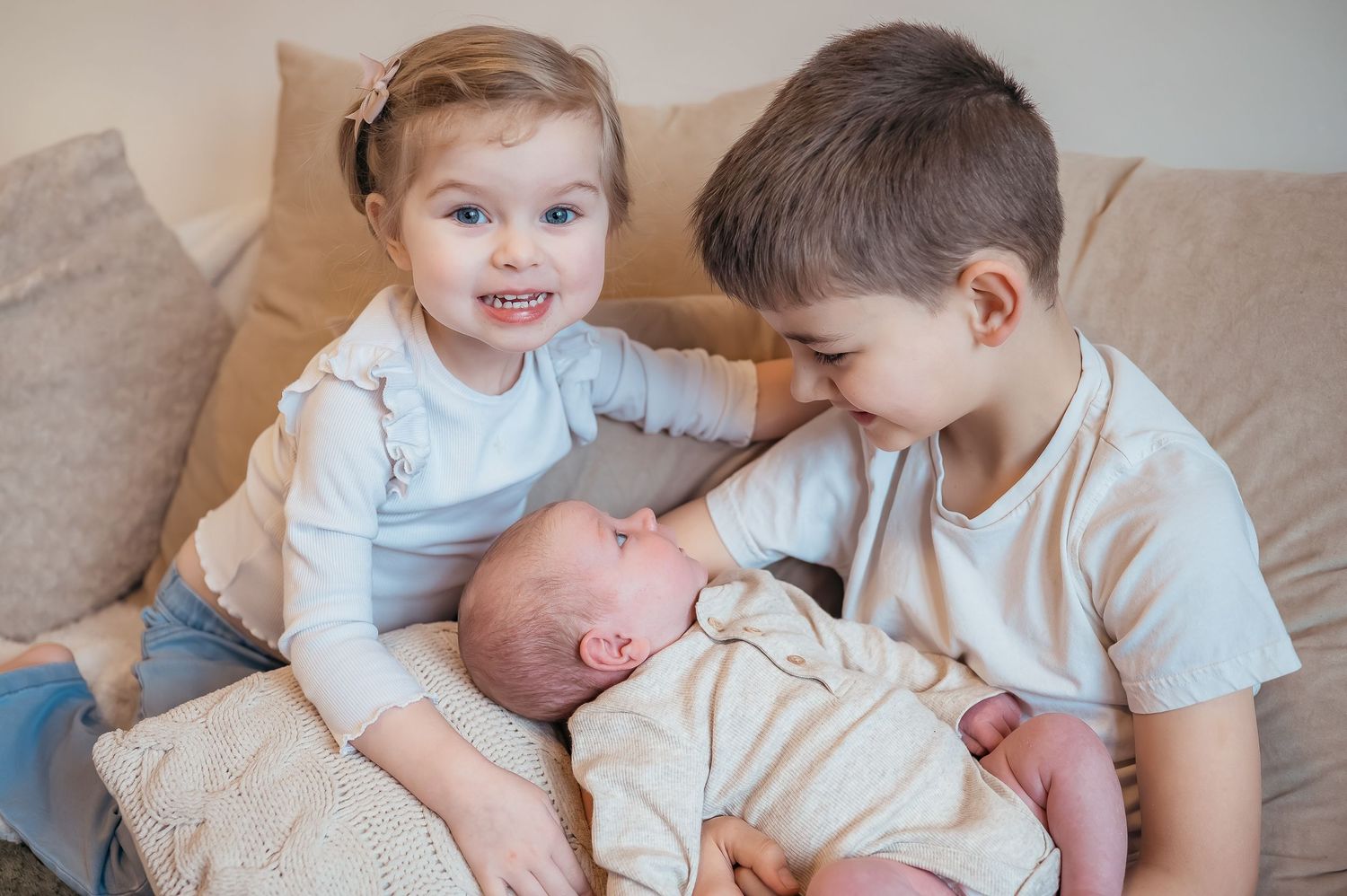 Two young siblings sitting on couch smiling while cradling sleeping newborn.