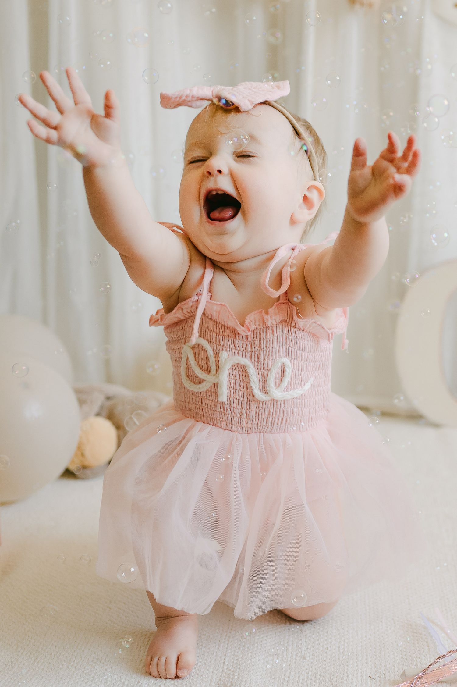 Baby girl in pink tutu dress celebrating first birthday with arms raised joyfully.