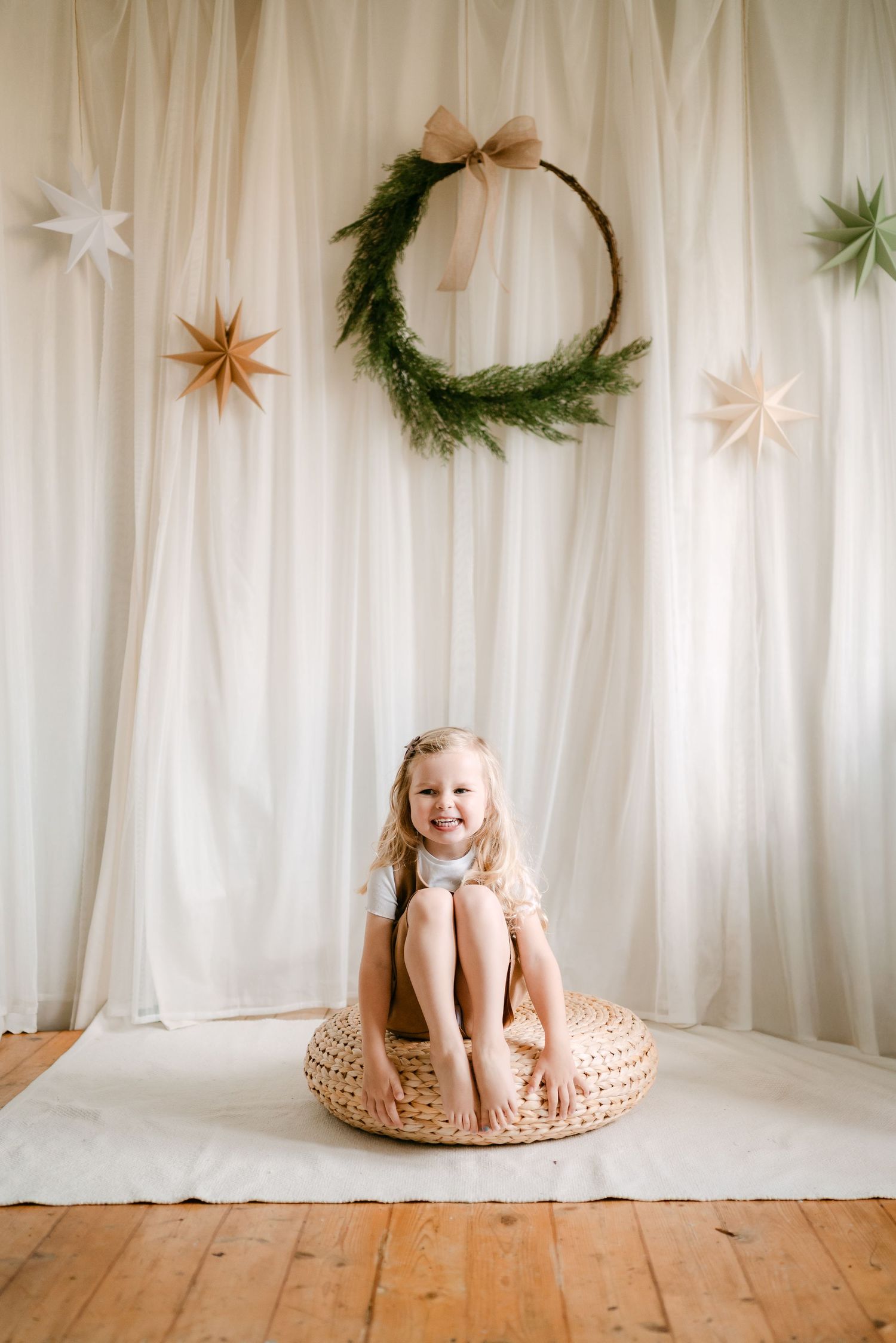 Smiling girl sitting on wicker pouf before white drape backdrop with green wreath and paper stars.