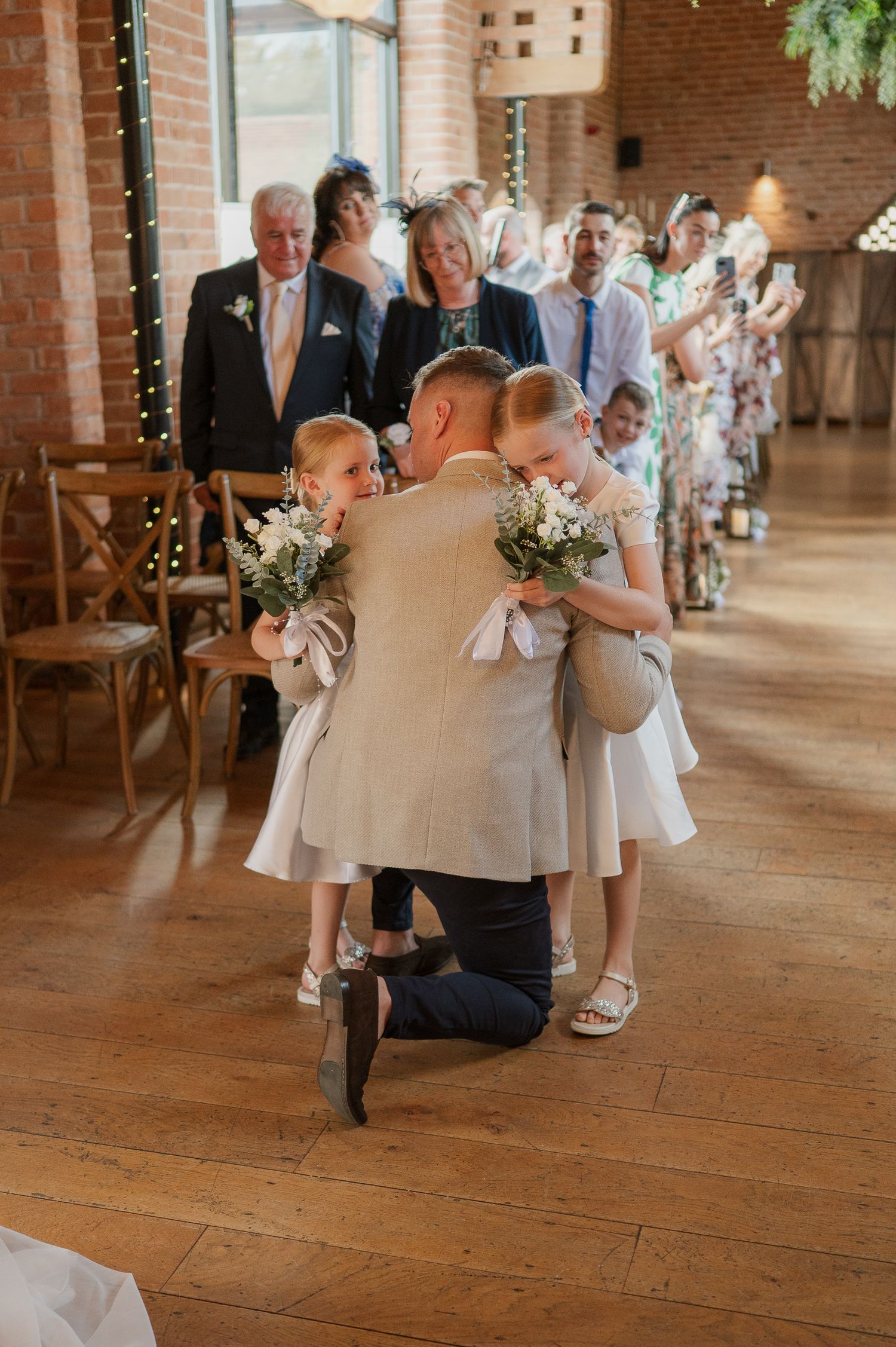 Groom kneels to embrace flower girls in white dresses during wedding ceremony aisle in rustic brick venue.