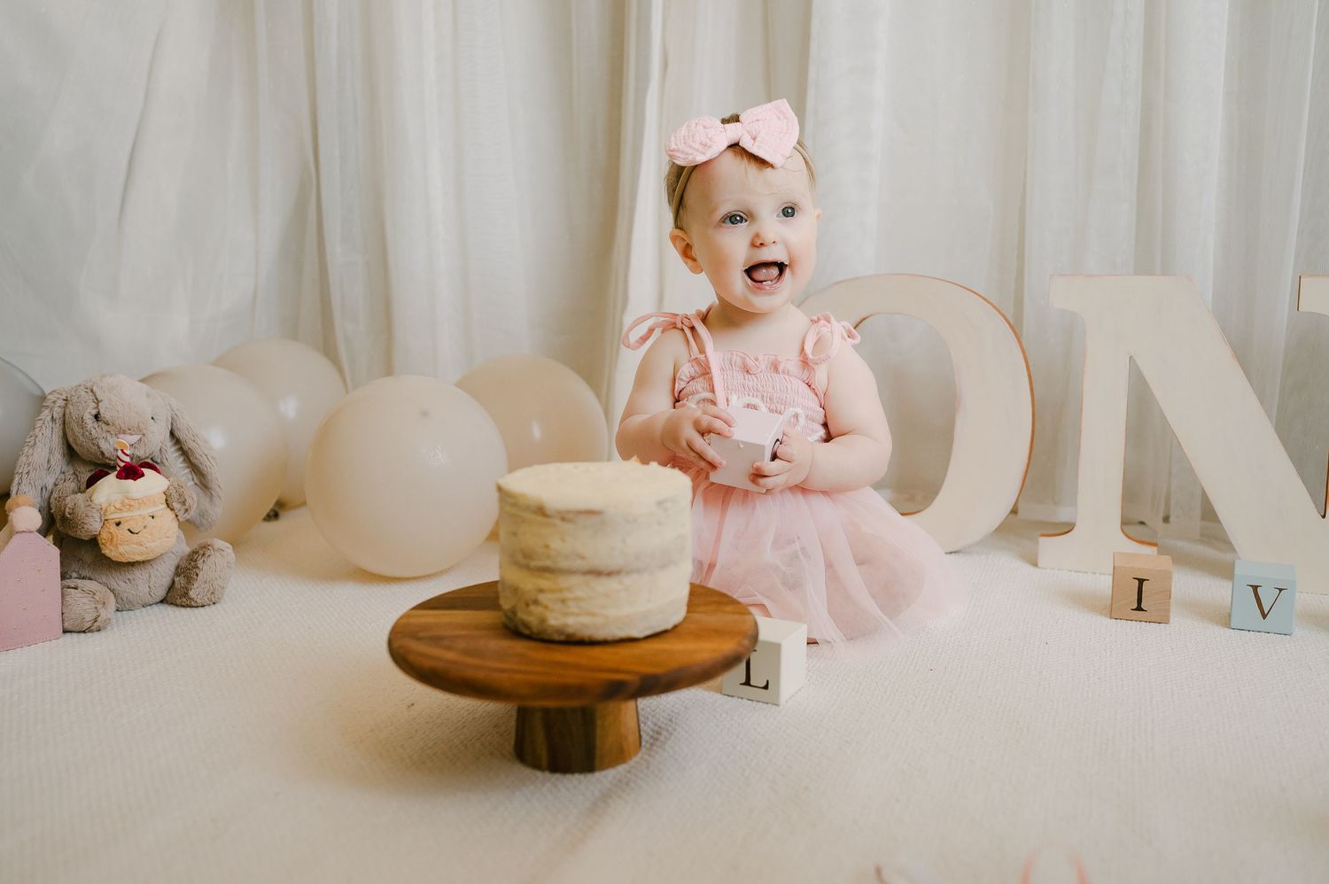 Smiling baby girl in pink dress with bow headband sitting before her first birthday cake.
