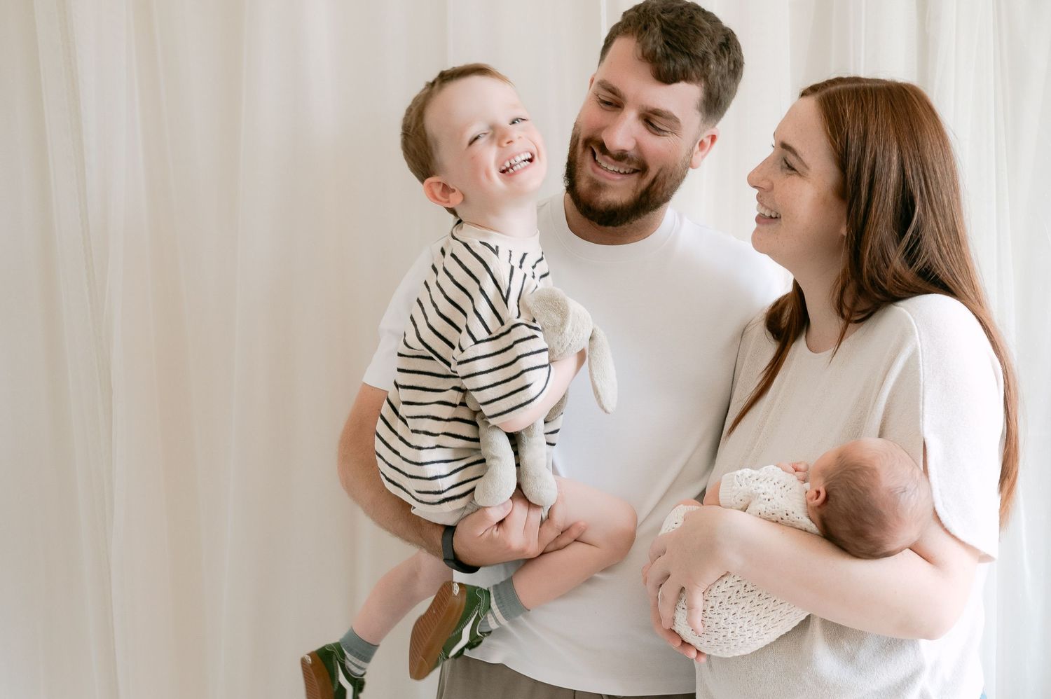 Smiling family with toddler and newborn baby posing against white backdrop.