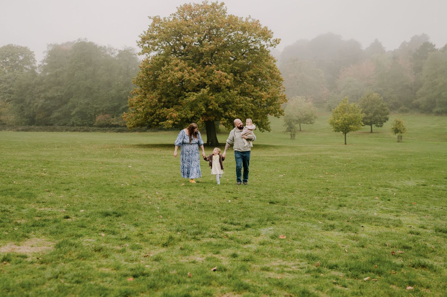 Family of four walking together on misty green field with autumn tree backdrop.