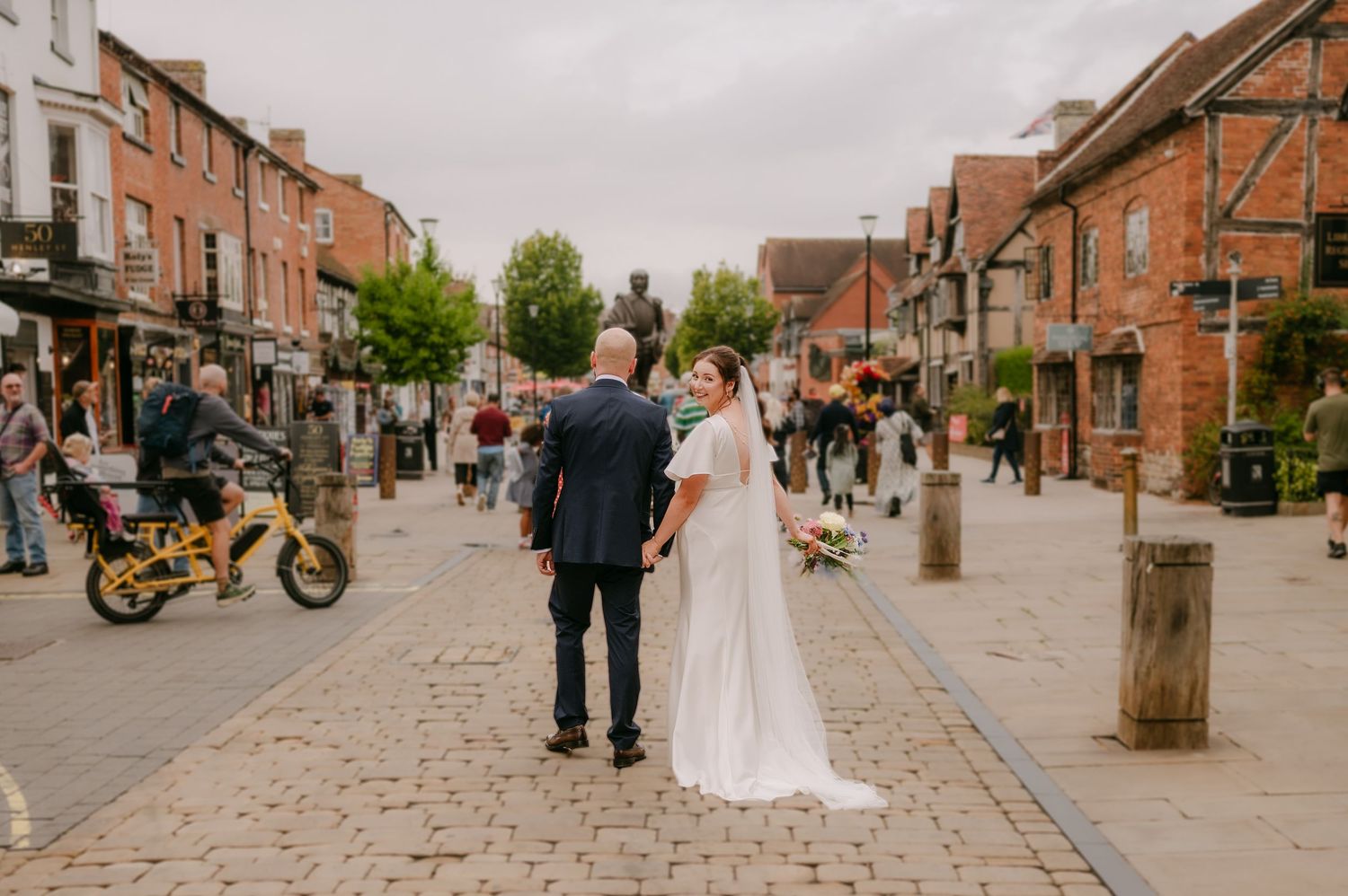 Bride and groom holding hands walking through a cobblestone UK town centre street surrounded by shops and pedestrians.