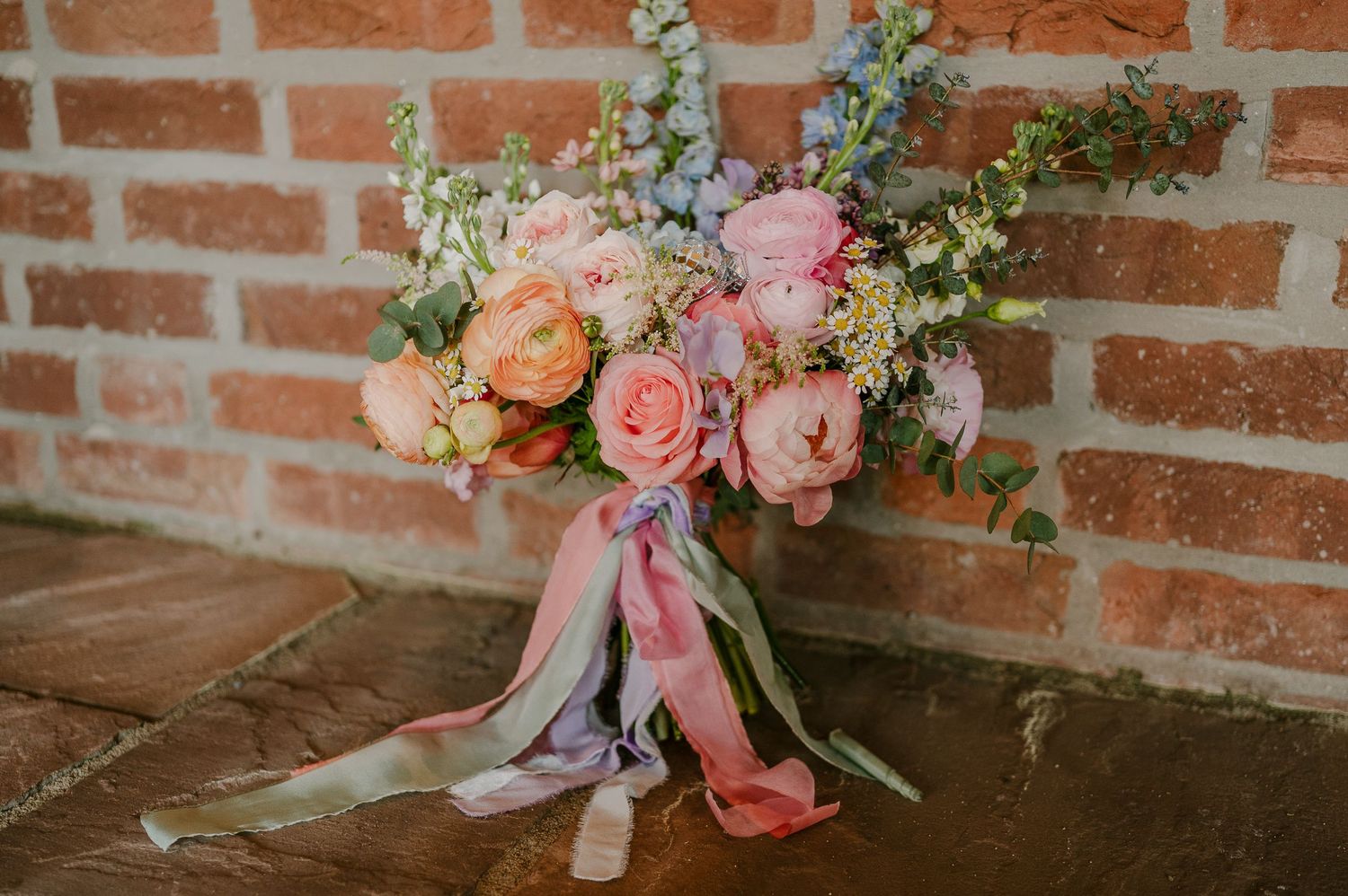 Romantic bridal bouquet of pink roses and pastel flowers with silk ribbons against rustic brick wall.