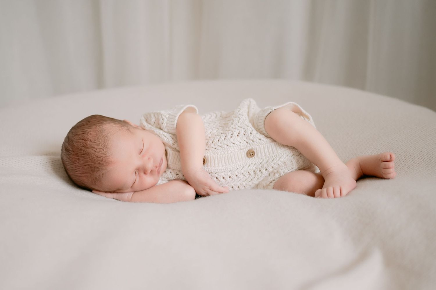 Sleeping newborn baby in cream knit romper curled on white blanket.