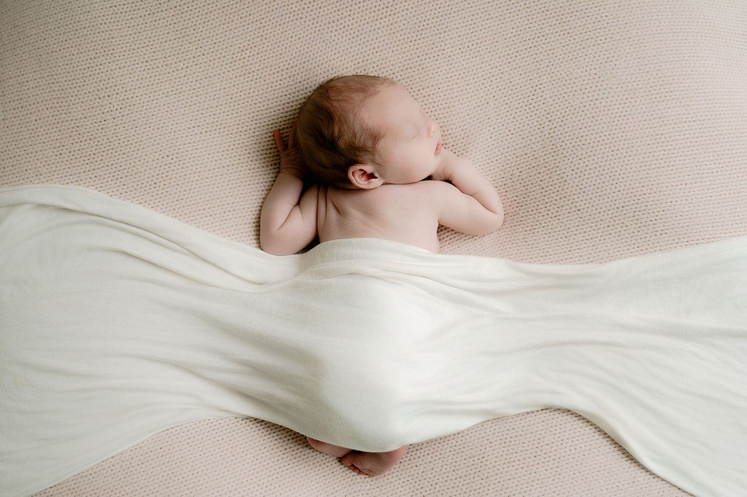 Overhead view of a sleeping newborn baby draped with a flowing white muslin wrap on cream background.