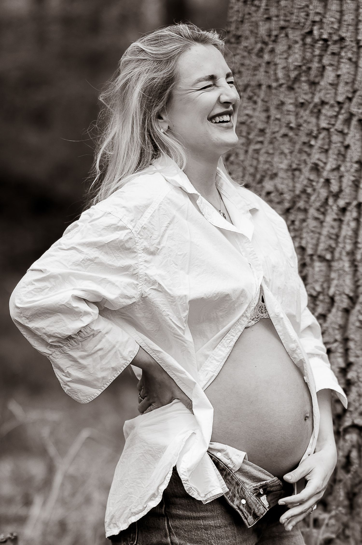 Black and white portrait of laughing pregnant woman in white shirt leaning against a tree.