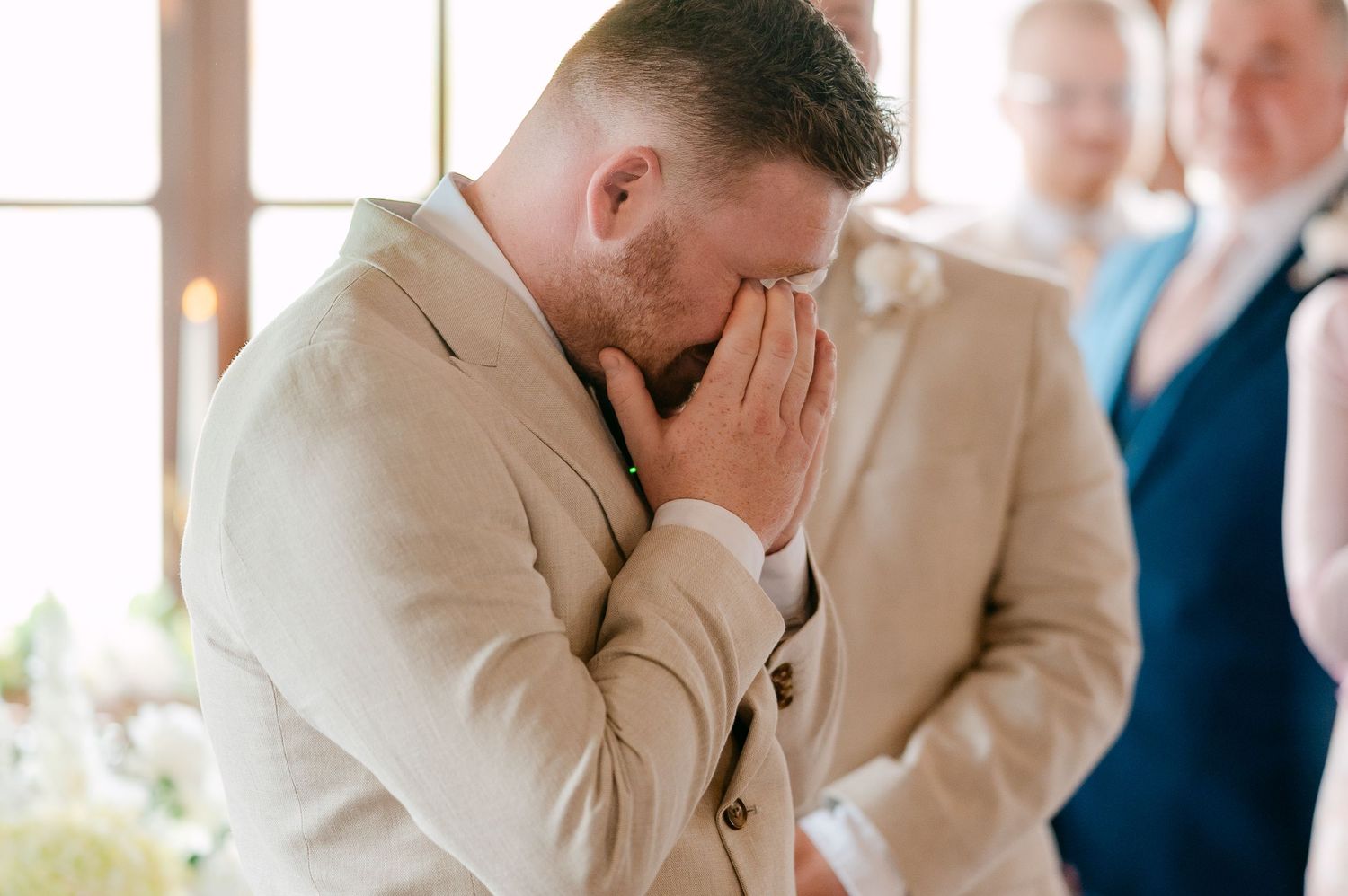Emotional groom in beige suit covers face with hands during heartfelt wedding ceremony moment.