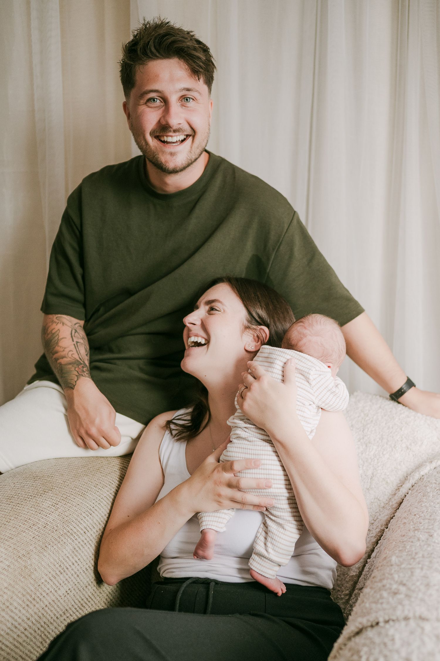 Happy new parents laughing together on couch holding their newborn baby.