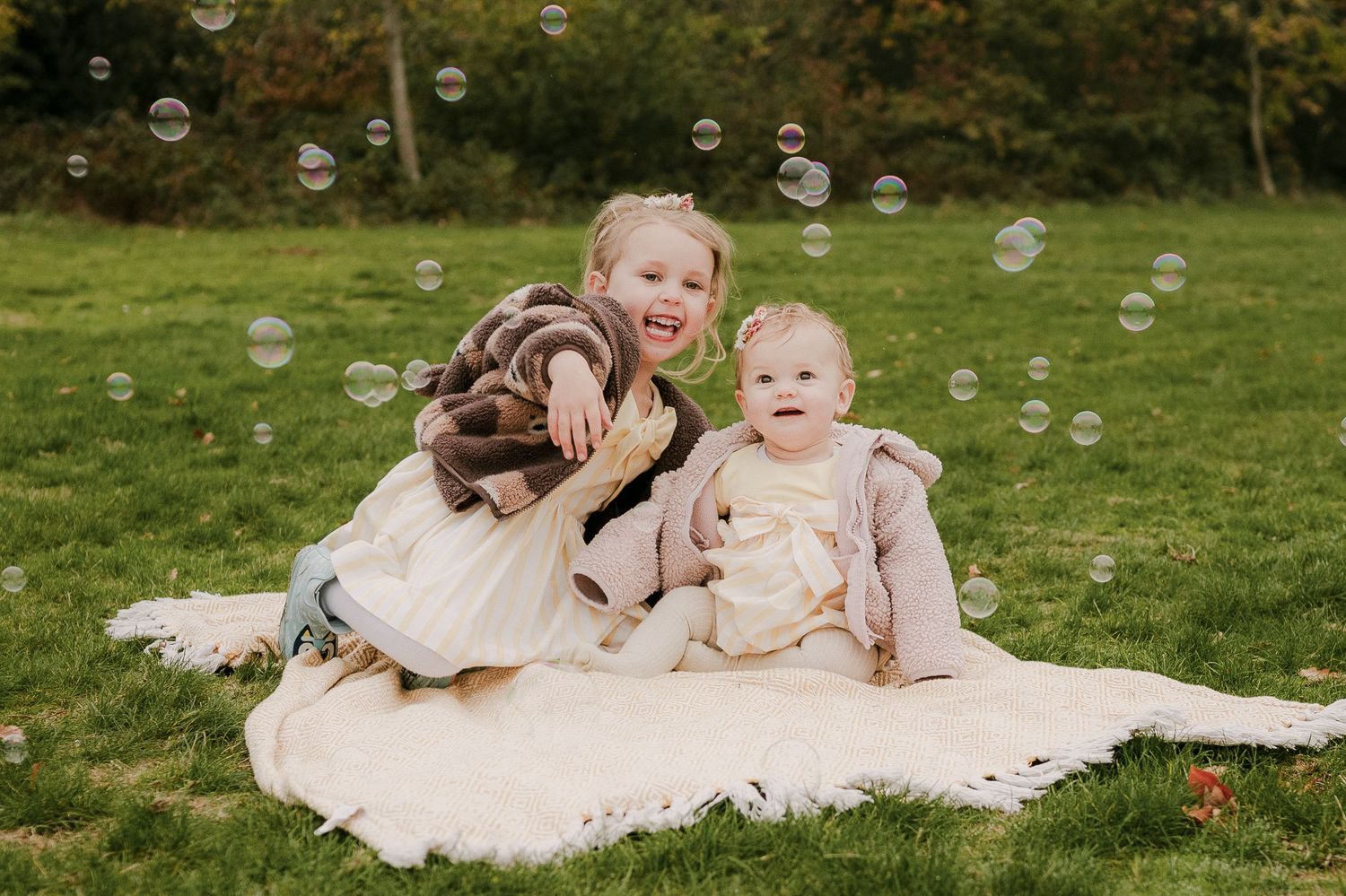 Two young siblings sitting on a blanket in a garden surrounded by floating soap bubbles.
