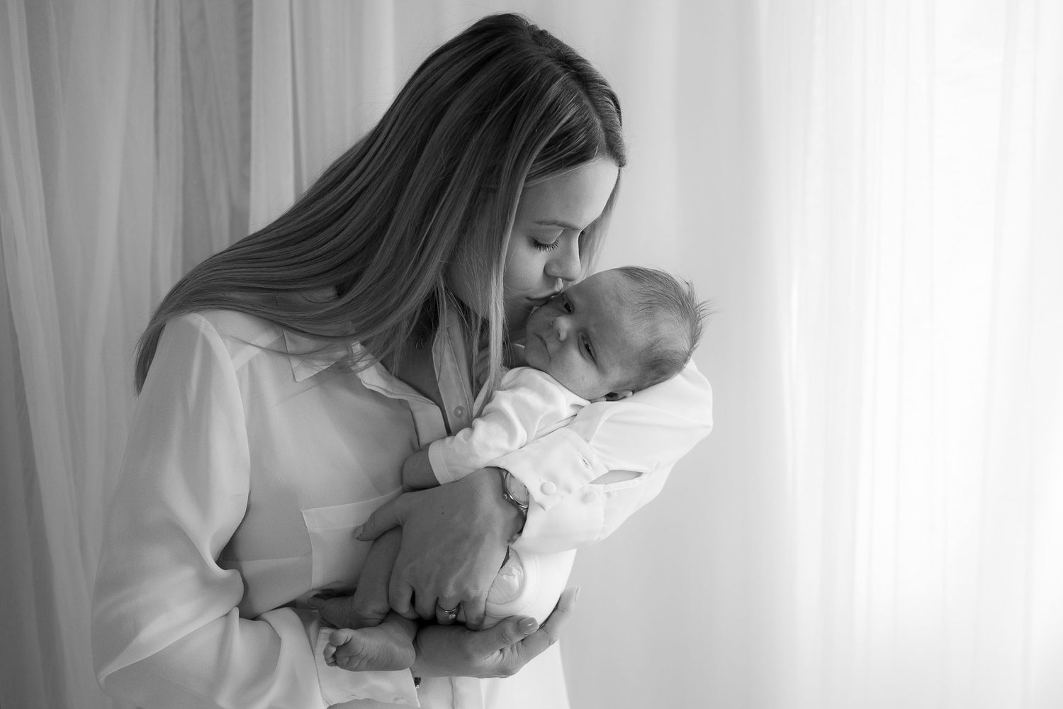 Black and white portrait of a mother tenderly kissing her sleeping newborn baby by a window.