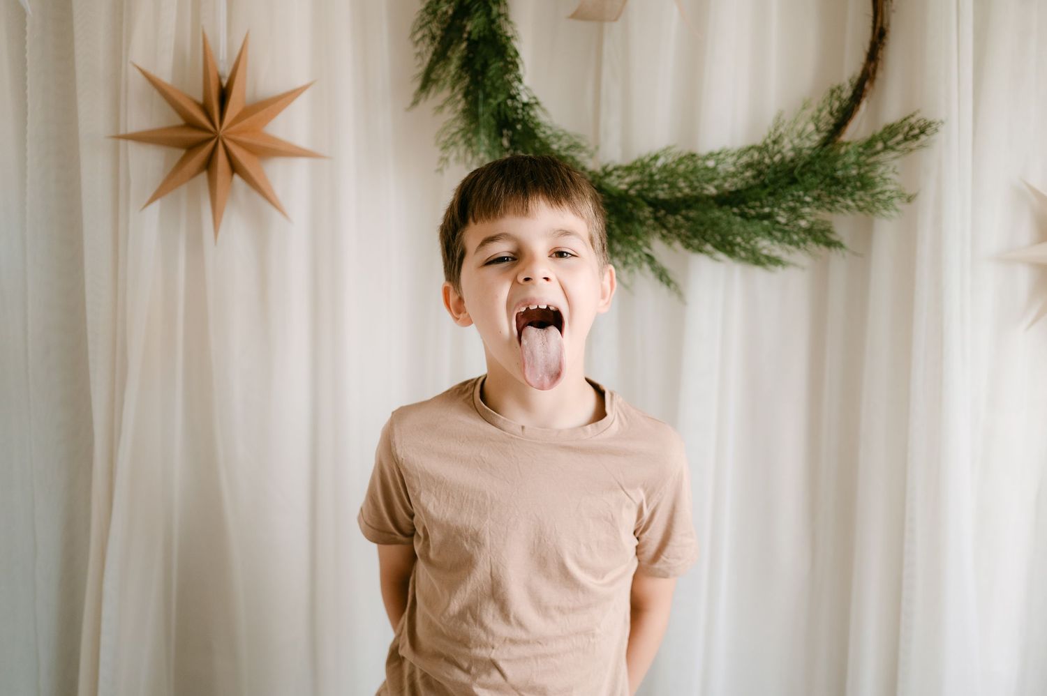 A young boy sticking his tongue out in front of a festive backdrop with a green wreath and star decoration.