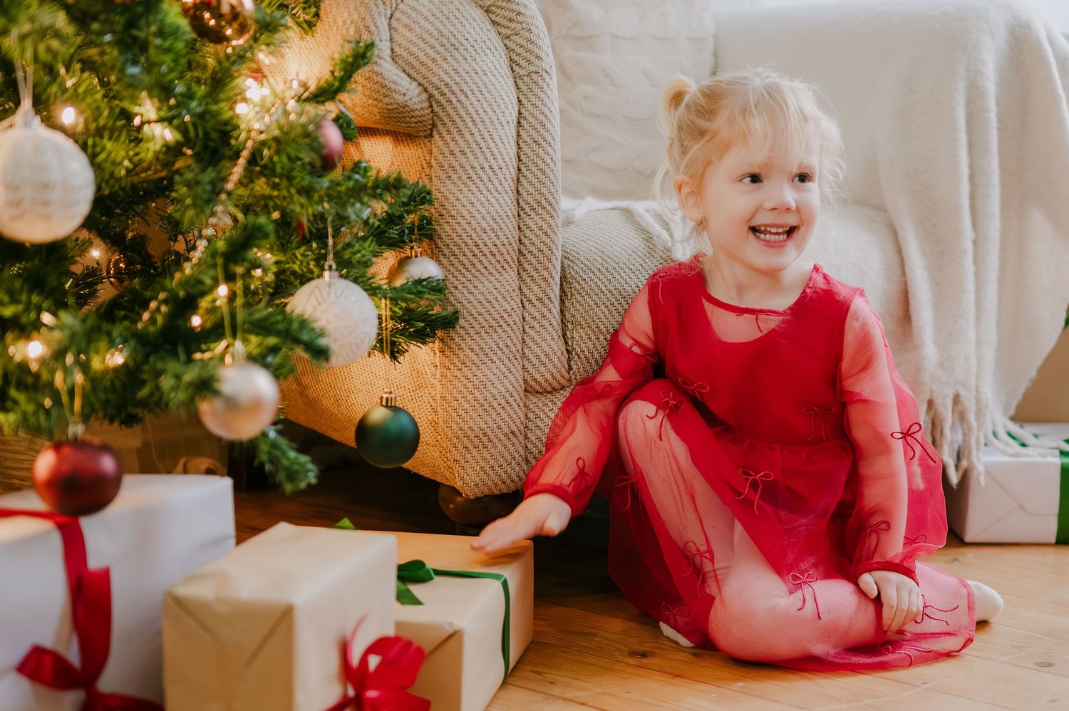 Cheerful toddler girl in red dress laughing while sitting beside Christmas tree surrounded by wrapped gifts.