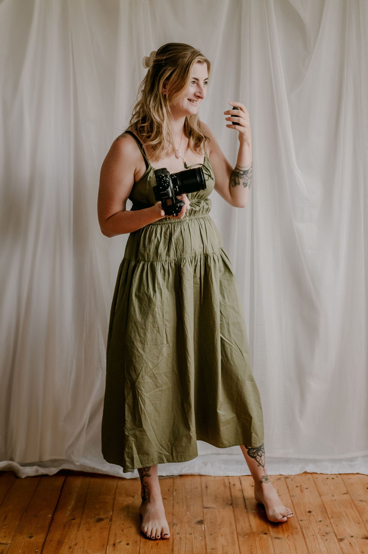 Smiling female photographer in olive green dress holding camera in bright white studio backdrop.