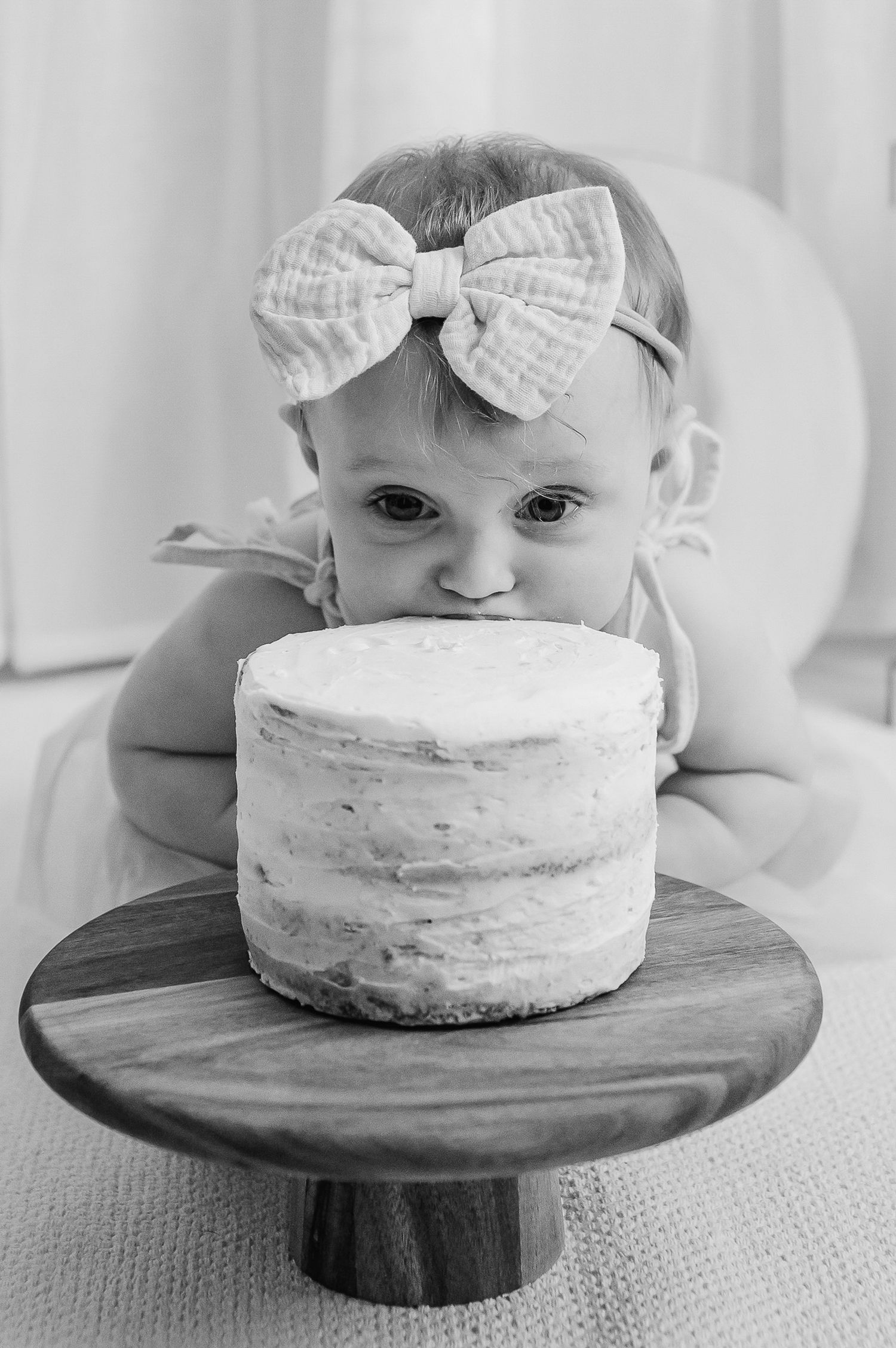 Black and white photo of baby girl in bow headband biting into a cake on a wooden stand.